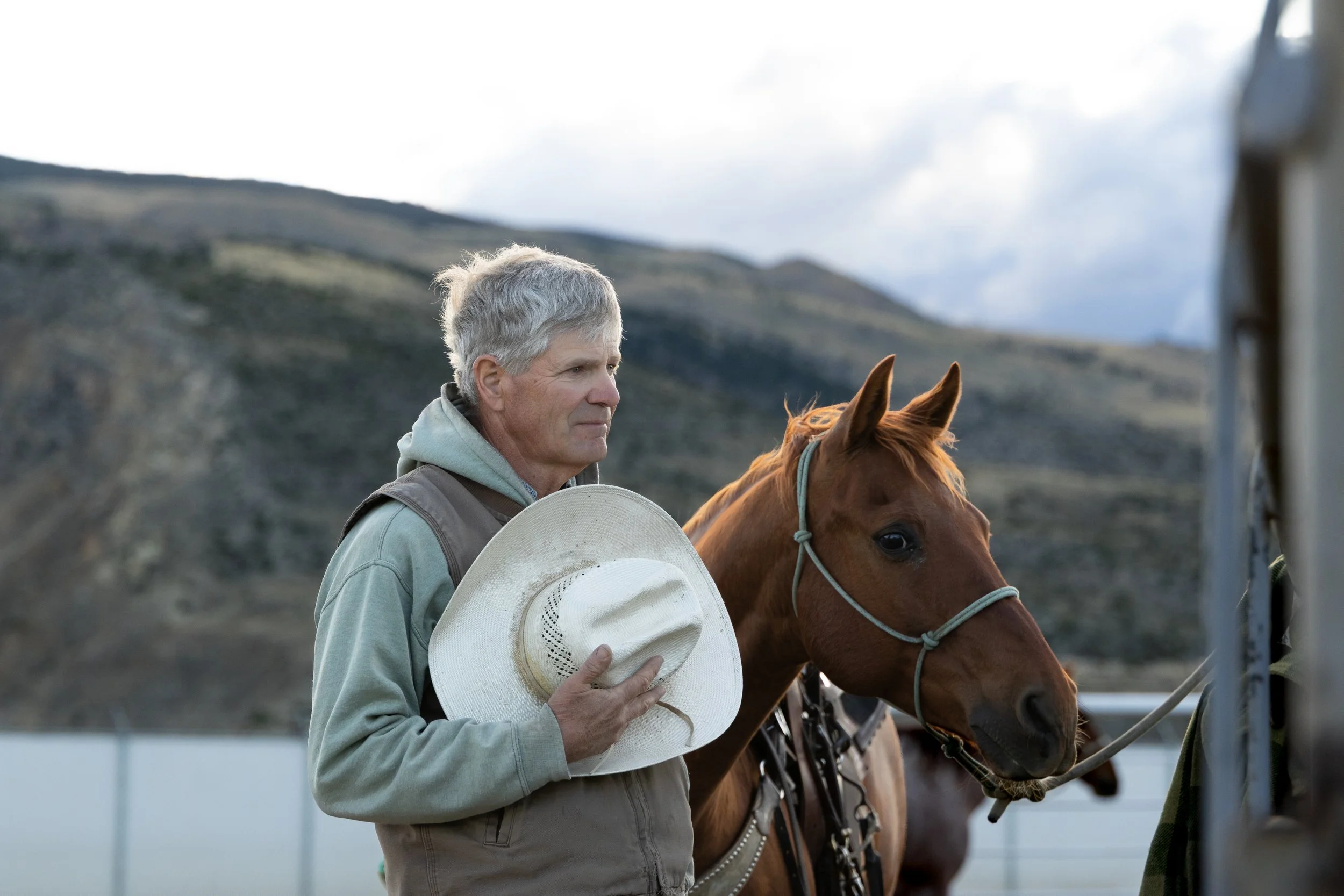 An older man with gray hair stands beside a brown horse, holding a white cowboy hat in his hand. The man is at a rodeo in Cody, Wyoming.