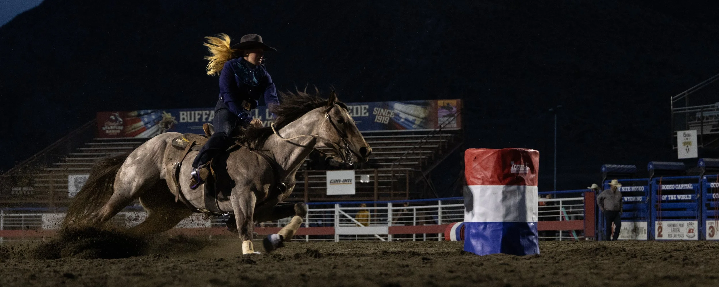 A woman riding a horse at night during a rodeo event, navigating around a red, white, and blue barrel.