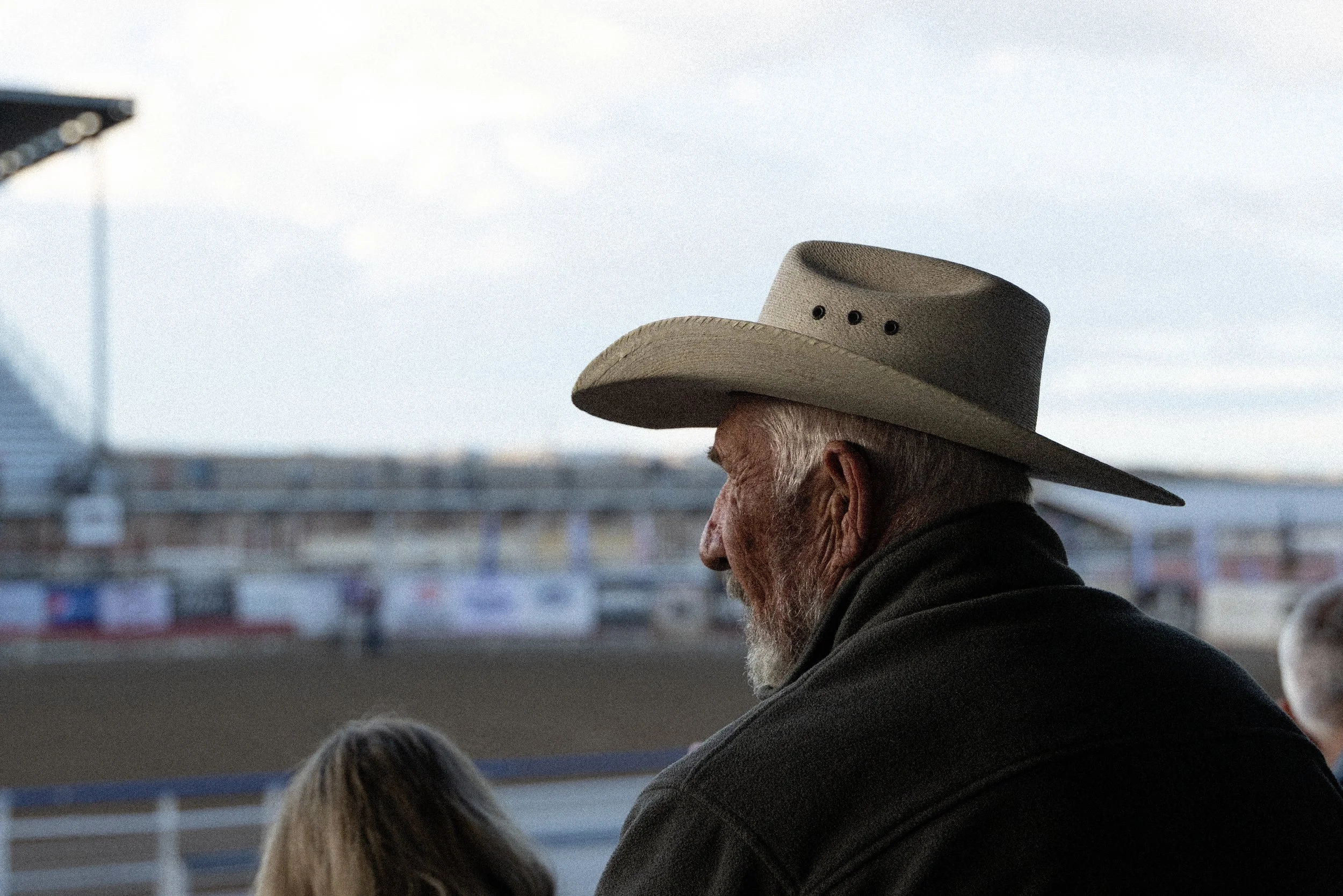 Profile of an elderly man with white hair and beard wearing a large beige cowboy hat, sitting outdoors at a location that looks like an amusement park or fairground, with blurred background structures and a cloudy sky.