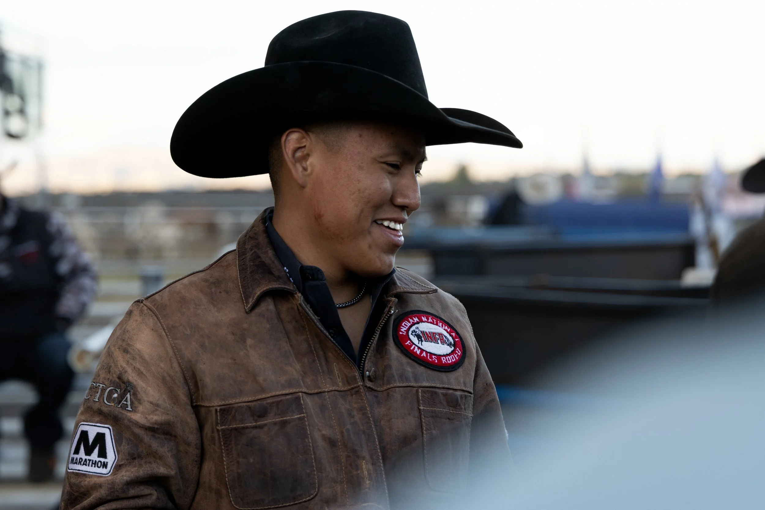 A person wearing a cowboy hat and a brown leather jacket with patches, smiling, standing outdoors near a marina or harbor with blurred boats and flags in the background.