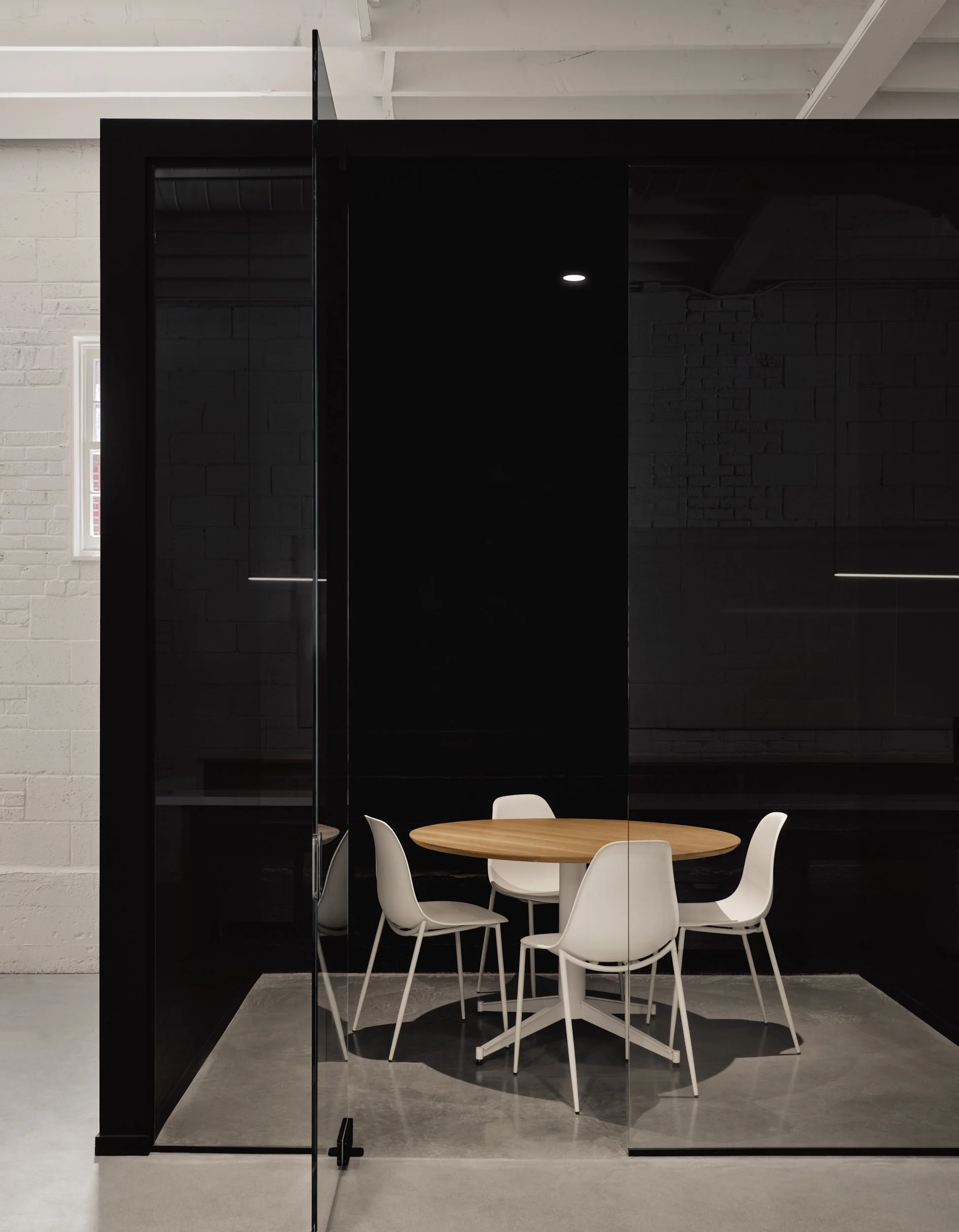 Salle de réunion avec un tableau rond en bois et six chaises blanches dans un espace moderne avec une cloison en verre et un mur noir.
