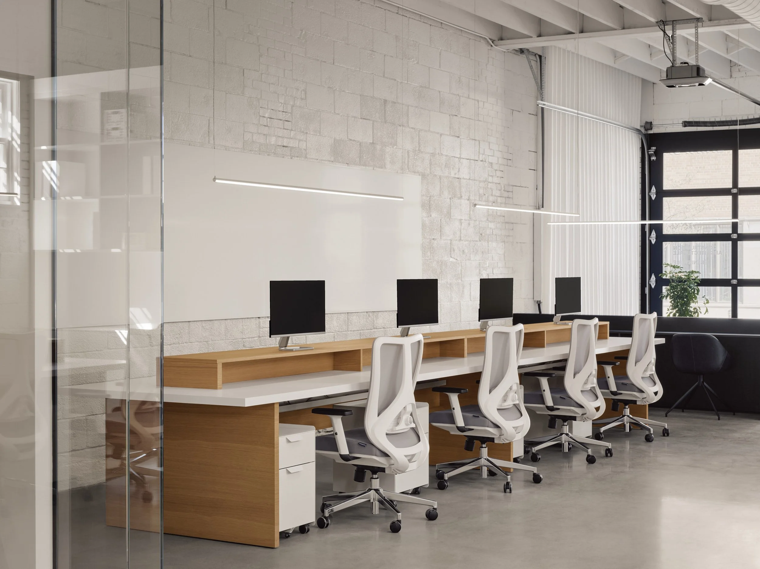 Bureau moderne avec quatre stations de travail, chaque station ayant un ordinateur, une chaise blanche ergonomique, dans un espace lumineux avec murs en briques blanches et grande fenêtre
