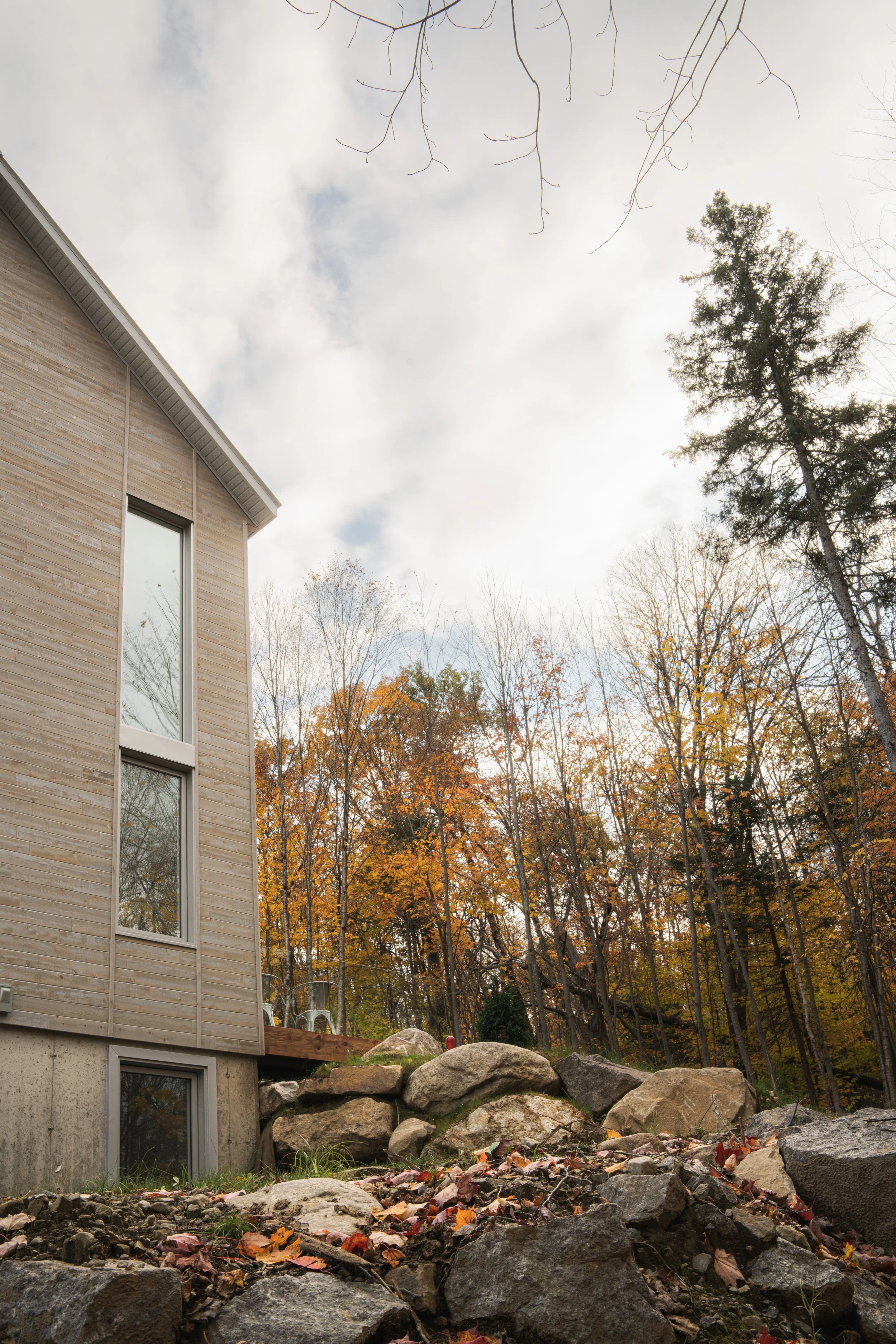 Extérieur d'une maison en bois avec de grandes fenêtres, entourée d'arbres aux feuilles d'automne, et des rochers dans la cour.