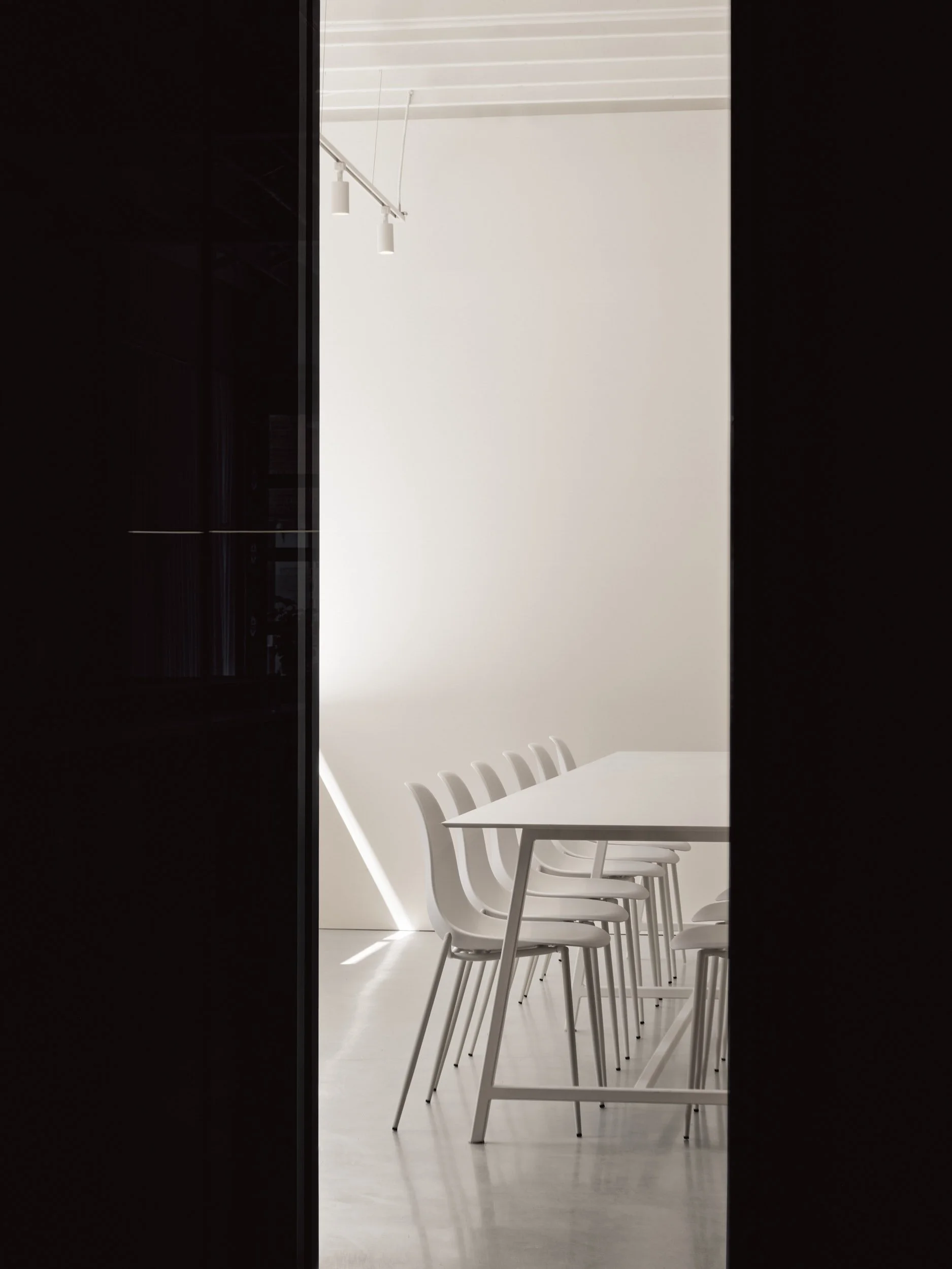 Salle de réunion moderne avec une longue table blanche et des chaises blanches, vue partielle à travers une porte ou une cloison sombre.