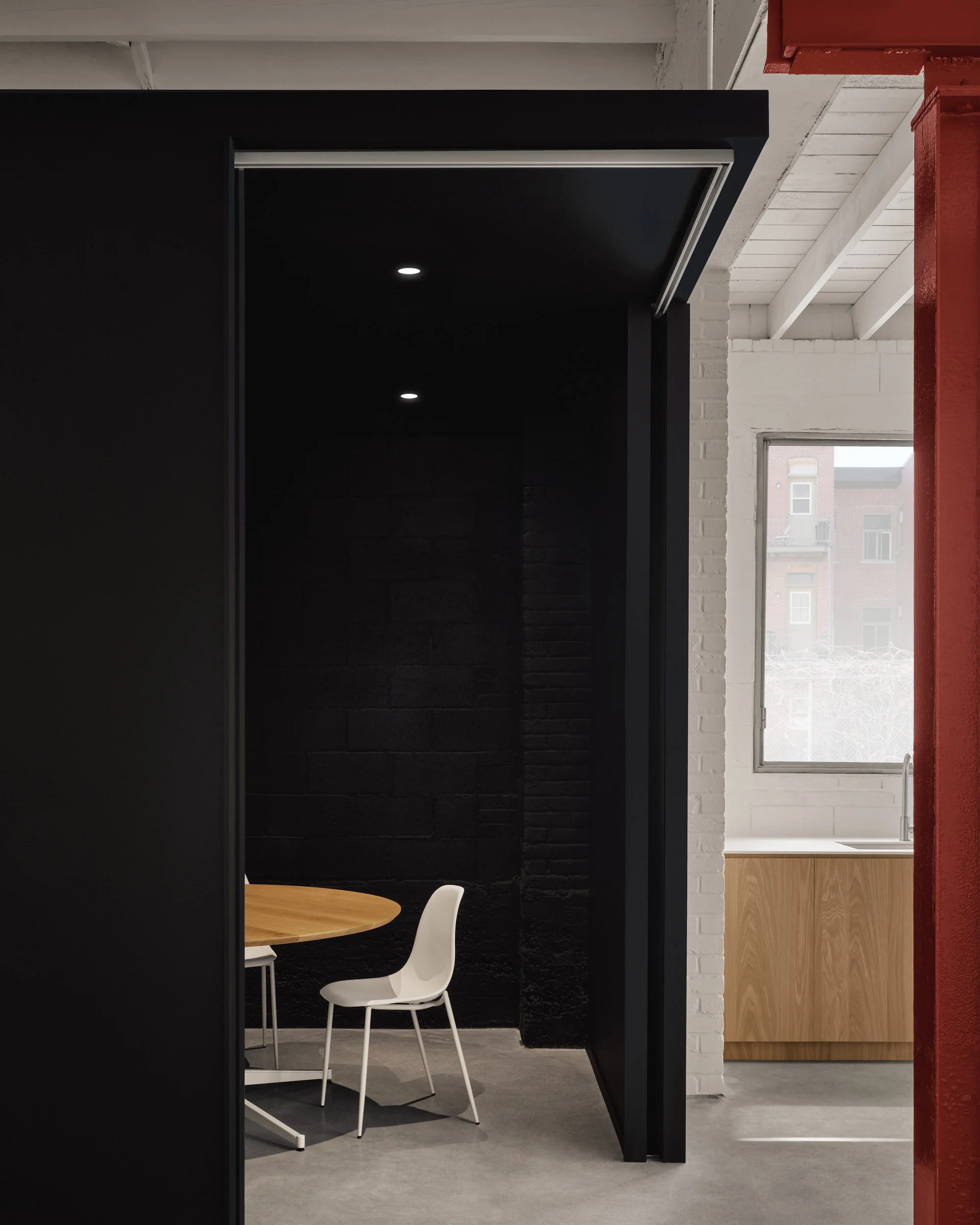Coin repas avec un mur noir, une table en bois ronde et une chaise blanche, à côté d'une cuisine avec un évier, une fenêtre et un mur en briques blanches.