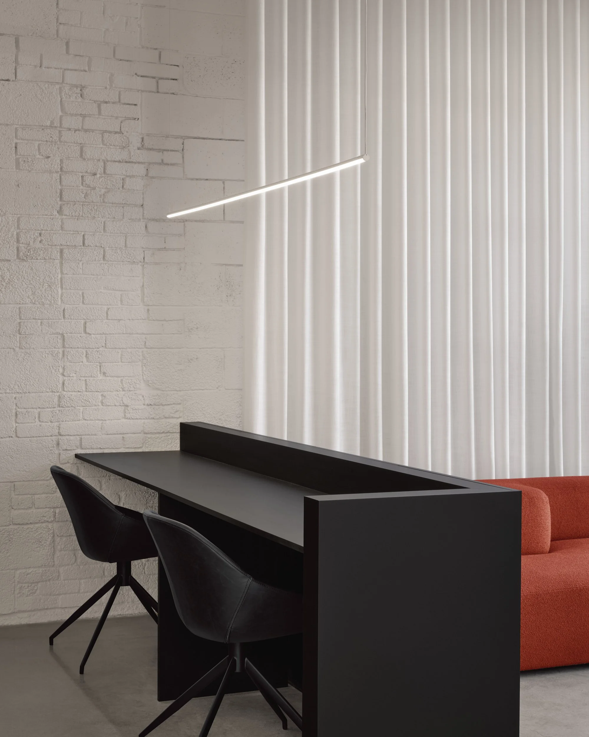 Espace de travail moderne avec un bureau noir, deux chaises noires, un canapé orange, un mur en brique blanc, des rideaux blancs et un éclairage suspendu.