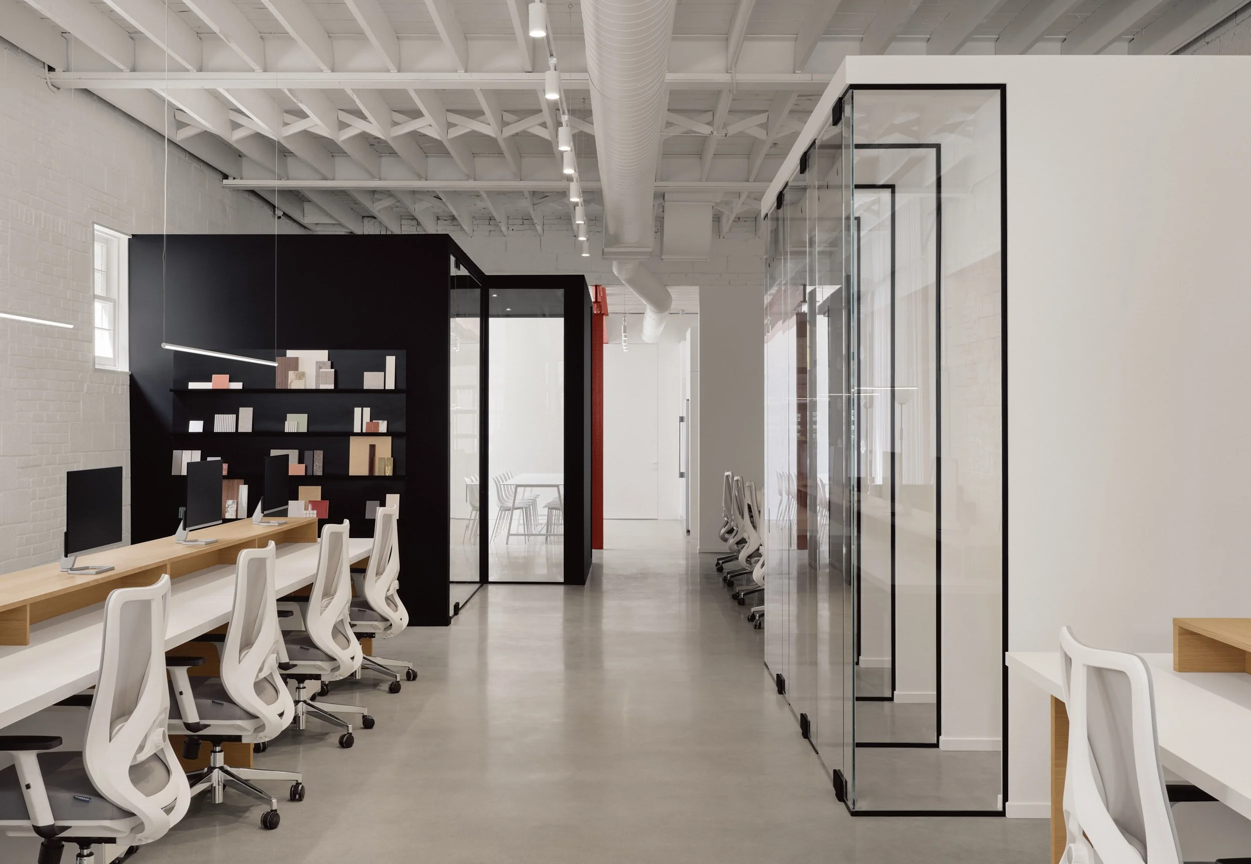 Espace de bureau moderne avec rangées de bureaux, chaises ergonomiques, ordinateurs, étagères noires avec livres, cloisons en verre et murs blancs, plafond industriel avec tuyauterie apparente.