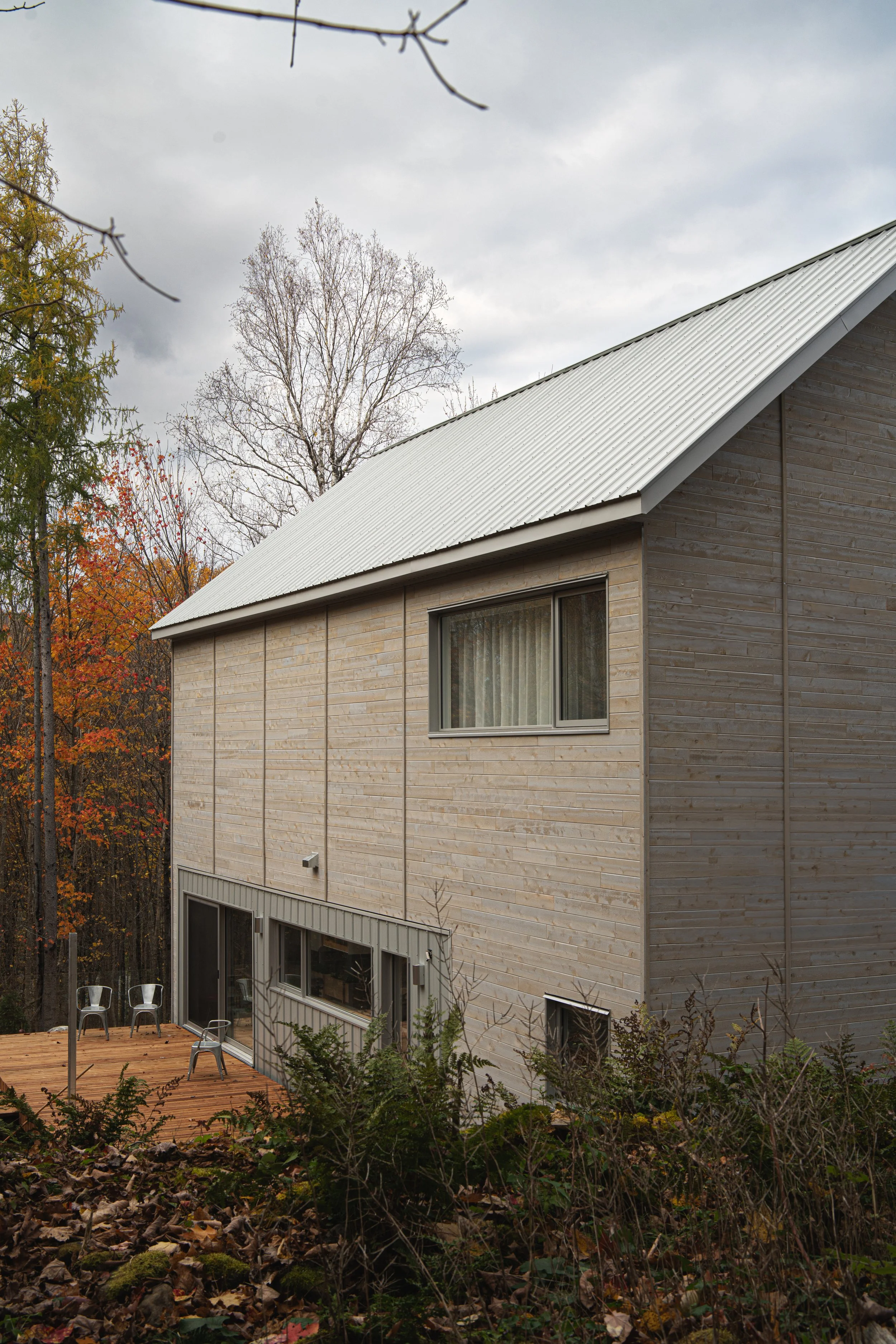 Maison en bois à deux étages avec terrasse en bois, fenêtres et toit en tôle, entourée de végétation automnale.