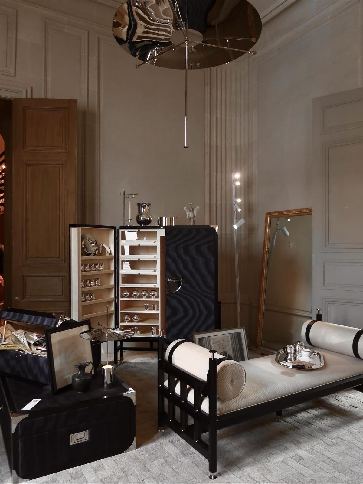 A room with a black and white modern sofa, a large mirror resting against the wall, and a display cabinet with glassware. The room has white paneled walls, a wooden wardrobe, and a metallic ceiling fan.