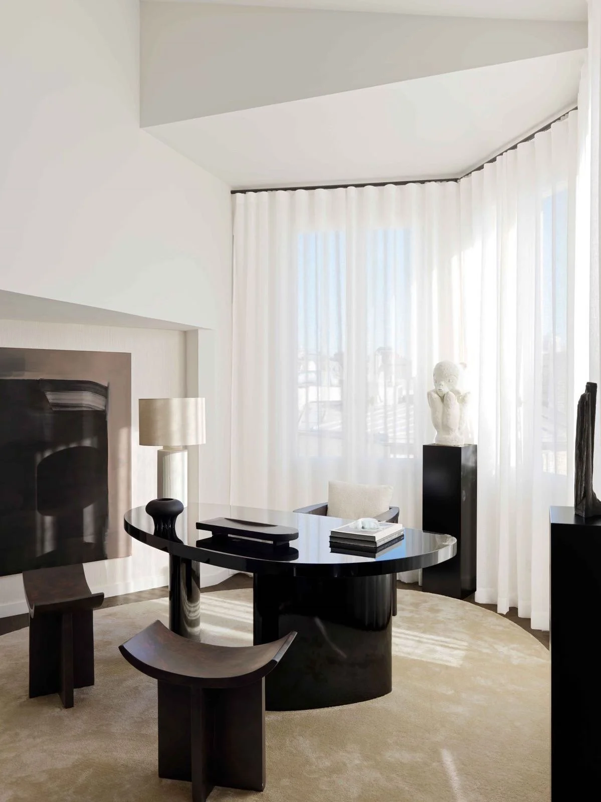 Modern interior with a black oval desk, two matching black stools, a white sculpture on a black pedestal, a table lamp, and white sheer curtains in a sunlit room.