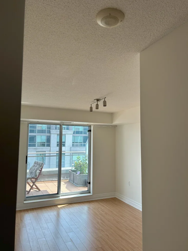 Empty room with hardwood flooring, sliding glass door leading to a balcony with outdoor chair and plant, ceiling light fixture, and a smoke detector.