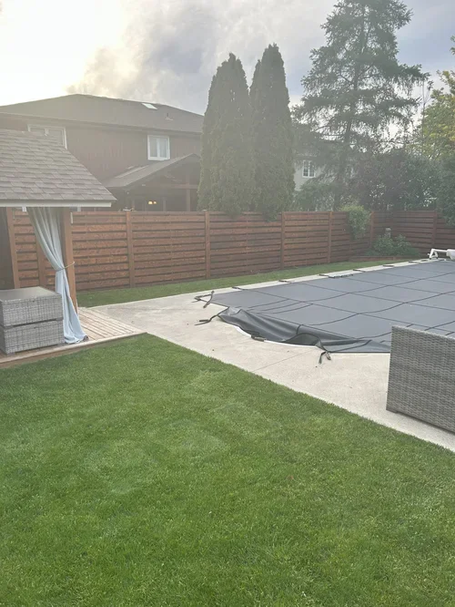Backyard with a swimming pool covered by a black pool cover, surrounded by a concrete patio and green grass, with a wooden fence and trees in the background, and a gazebo with outdoor furniture on the left.