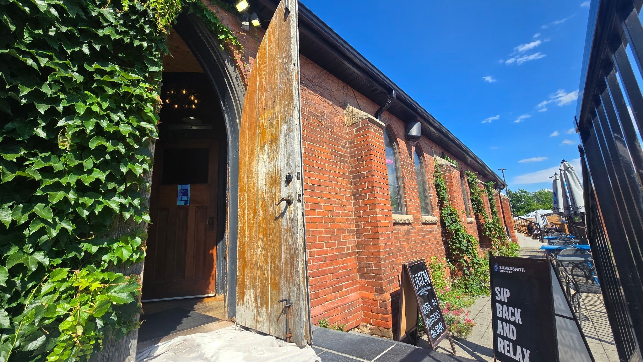 Brick building with arched windows, ivy on the wall, an open wooden door, and outdoor seating with umbrellas, signs advertising drinks and tagline 'Sip Back and Relax'.
