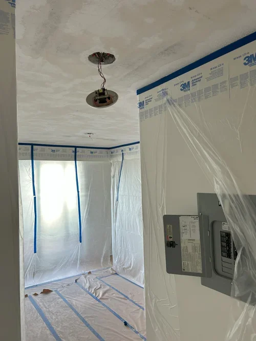 Room under construction with drywall and plastic sheeting covering windows and walls, patches for ceiling light fixture, and an electrical panel visible.