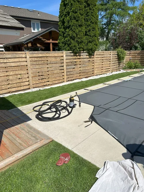 Backyard with wooden fence, green grass, trees, covered pool with cover, pool tools, and a pair of red clogs on the grass.