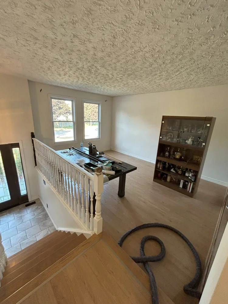 Living room with two windows, a staircase, a table with various objects, a glass cabinet, and a vacuum hose on the floor.