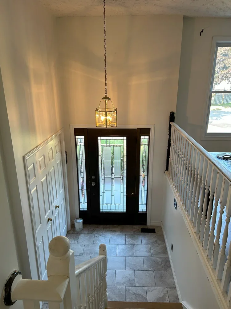 Interior view of a house entrance with a black front door with glass panels, a hanging ceiling light, a window on the right, a staircase with white railing, and tiled flooring.