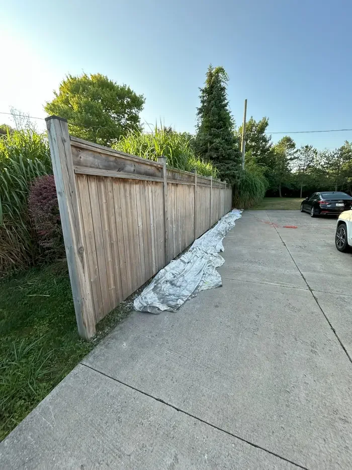 Wooden privacy fence with a tarp or fabric at the base, next to a parking lot with parked cars, green trees, and clear blue sky.