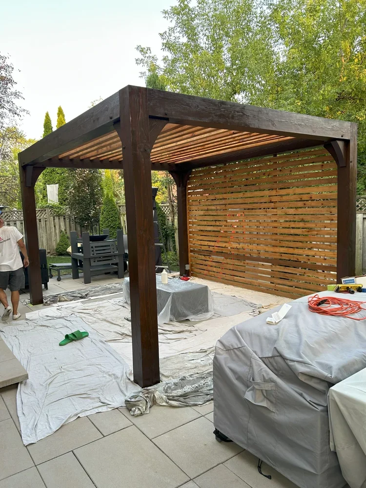 A backyard patio with a newly built wooden pergola, covered with tarps and protection sheets for finishing, surrounded by trees and outdoor furniture.