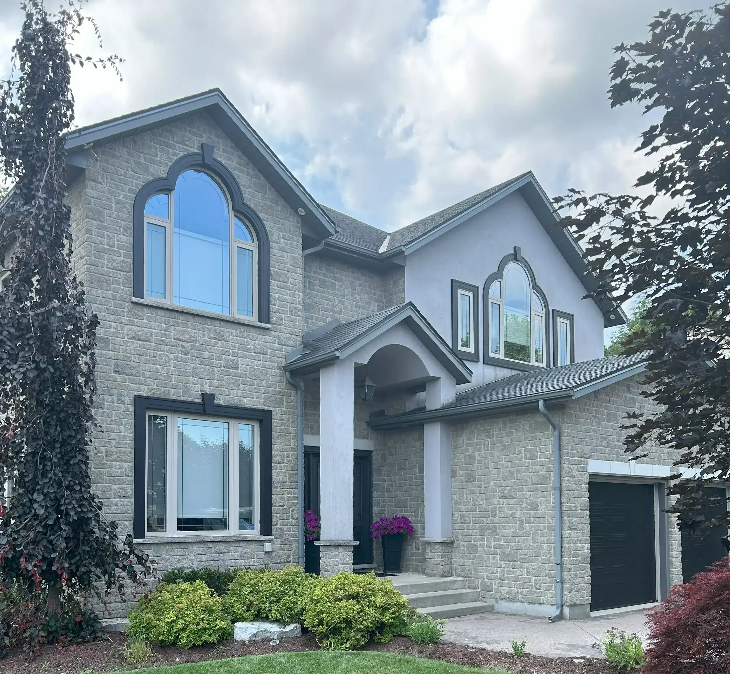 A two-story house with stone and stucco exterior, arched and rectangular windows, and a front porch with potted purple flowers, surrounded by green lawn and trees.