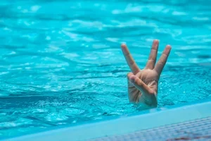 A person's hand making a three-finger peace sign emerging from the water in a swimming pool.