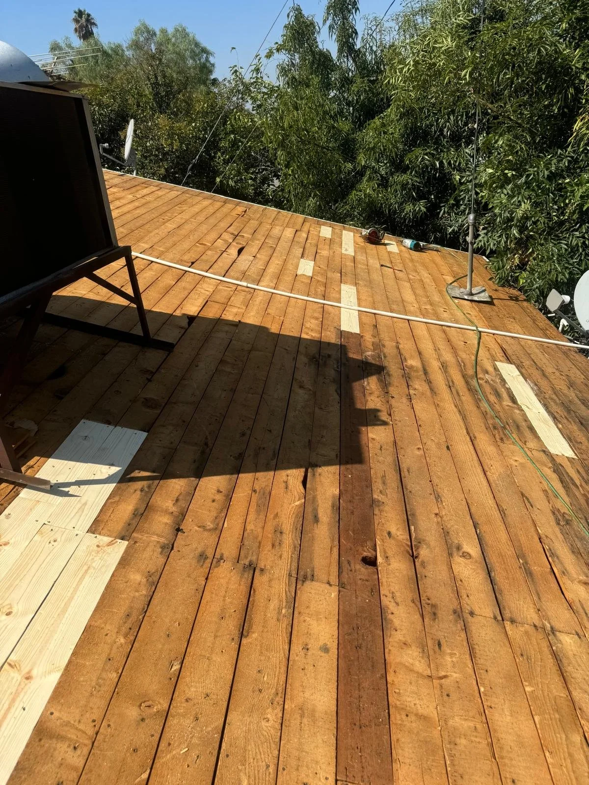 A wooden deck under construction with some unfinished planks and tools on the surface, surrounded by trees and outdoor furniture, with electrical wires overhead.