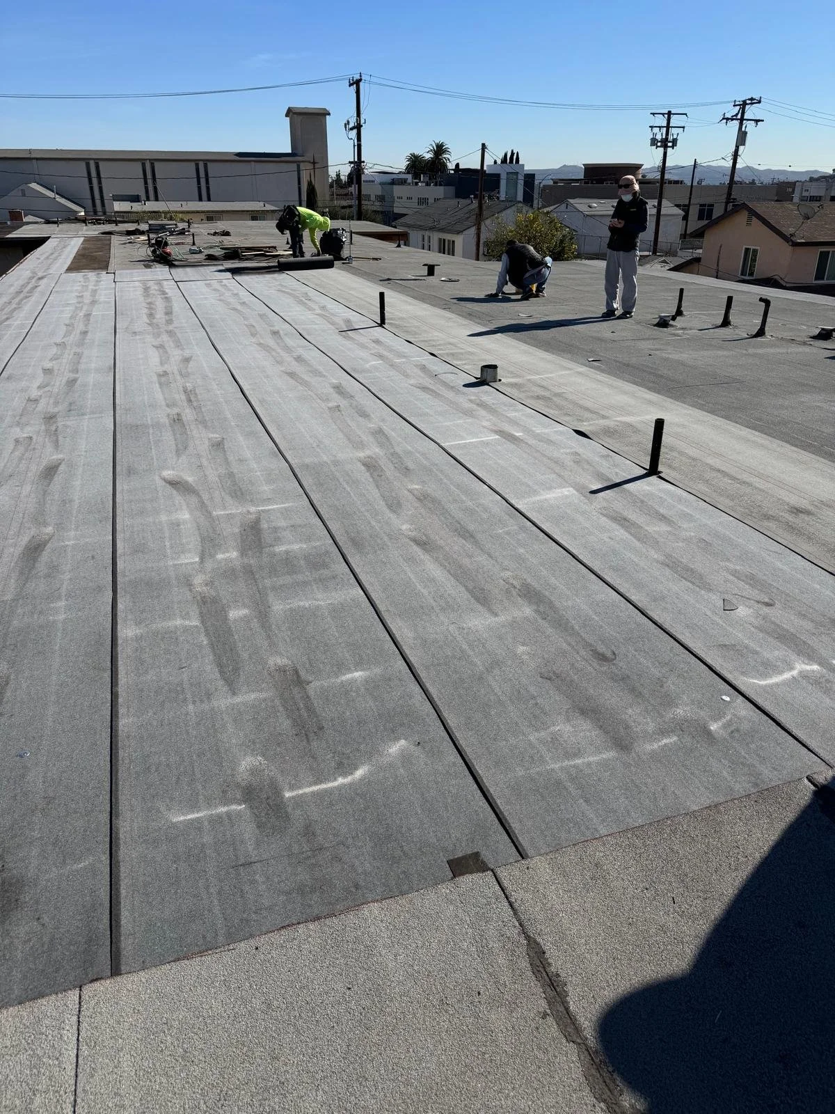Roof workers installing or repairing a flat rooftop with gray roofing material under a clear blue sky, with houses and power lines in the background.