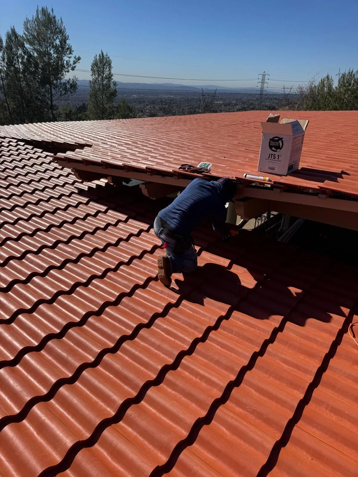 A person working on installing or repairing a red tiled roof, kneeling on the roof with tools and materials nearby, including a cardboard box and a small electronic device, with a view of distant trees and power lines under a blue sky.