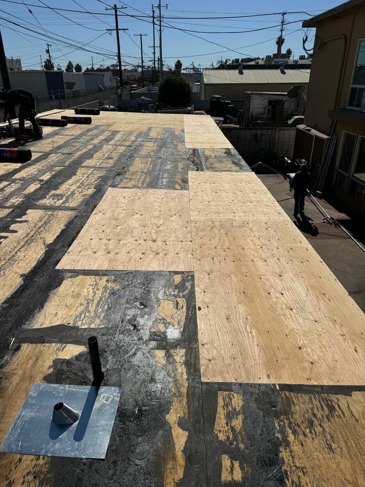 Roof under construction with plywood sheets and black waterproof membrane, with workers and equipment at the edge, under a clear sky with power lines overhead.