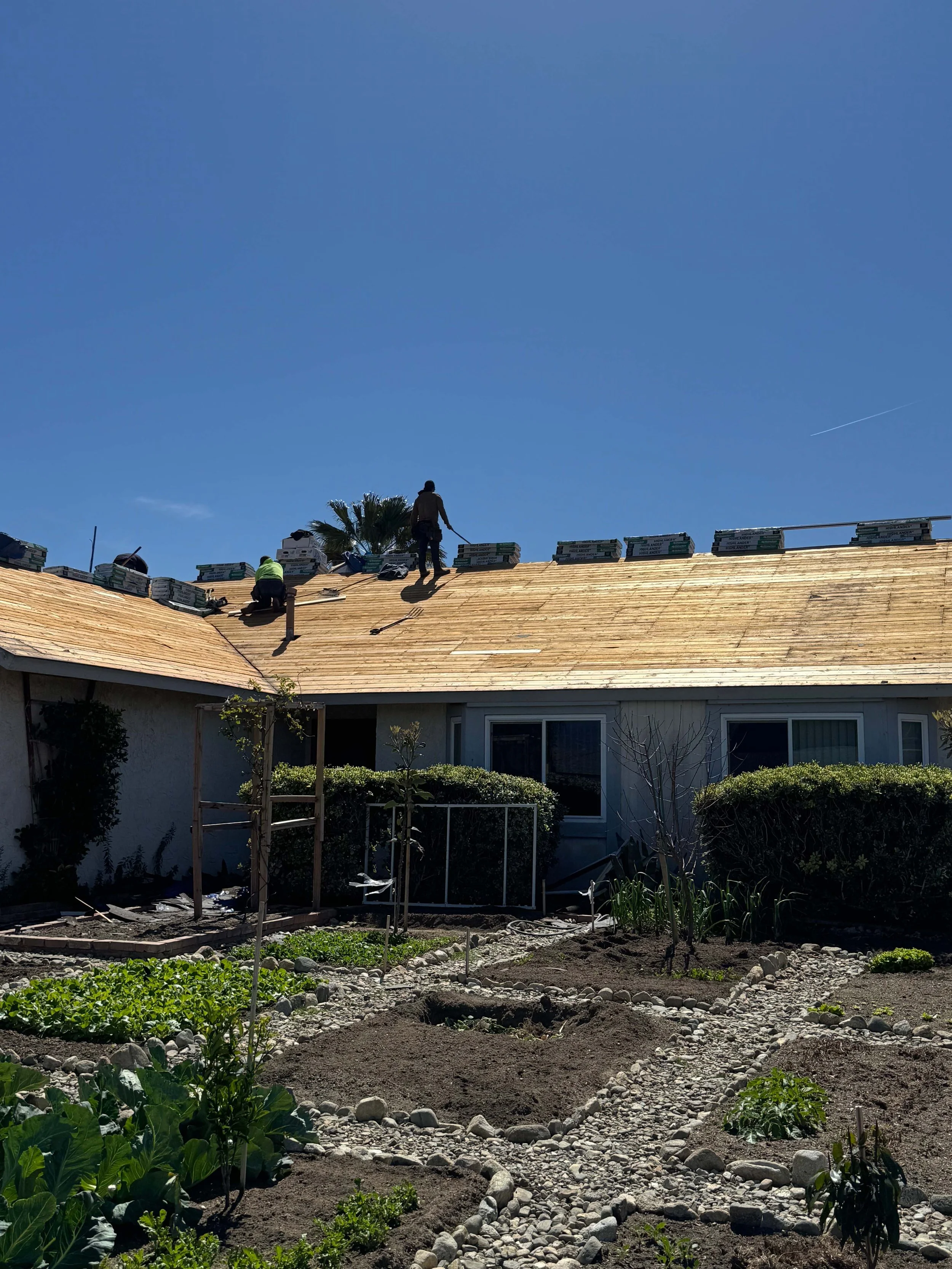 Construction workers on a house roof, with a garden in the foreground and a clear blue sky.