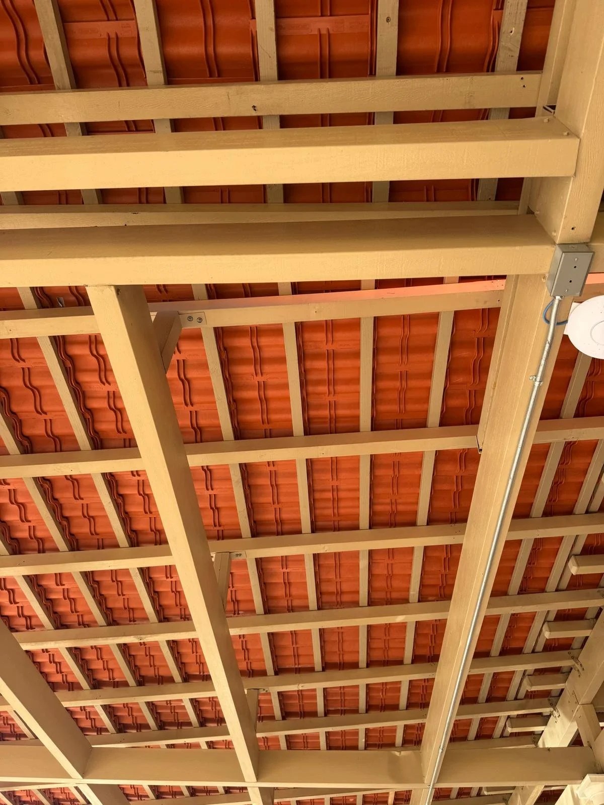 The photo shows a view of a wooden ceiling framework with a red tiled roof above it. There is a light fixture with a round bulb in the lower right corner.