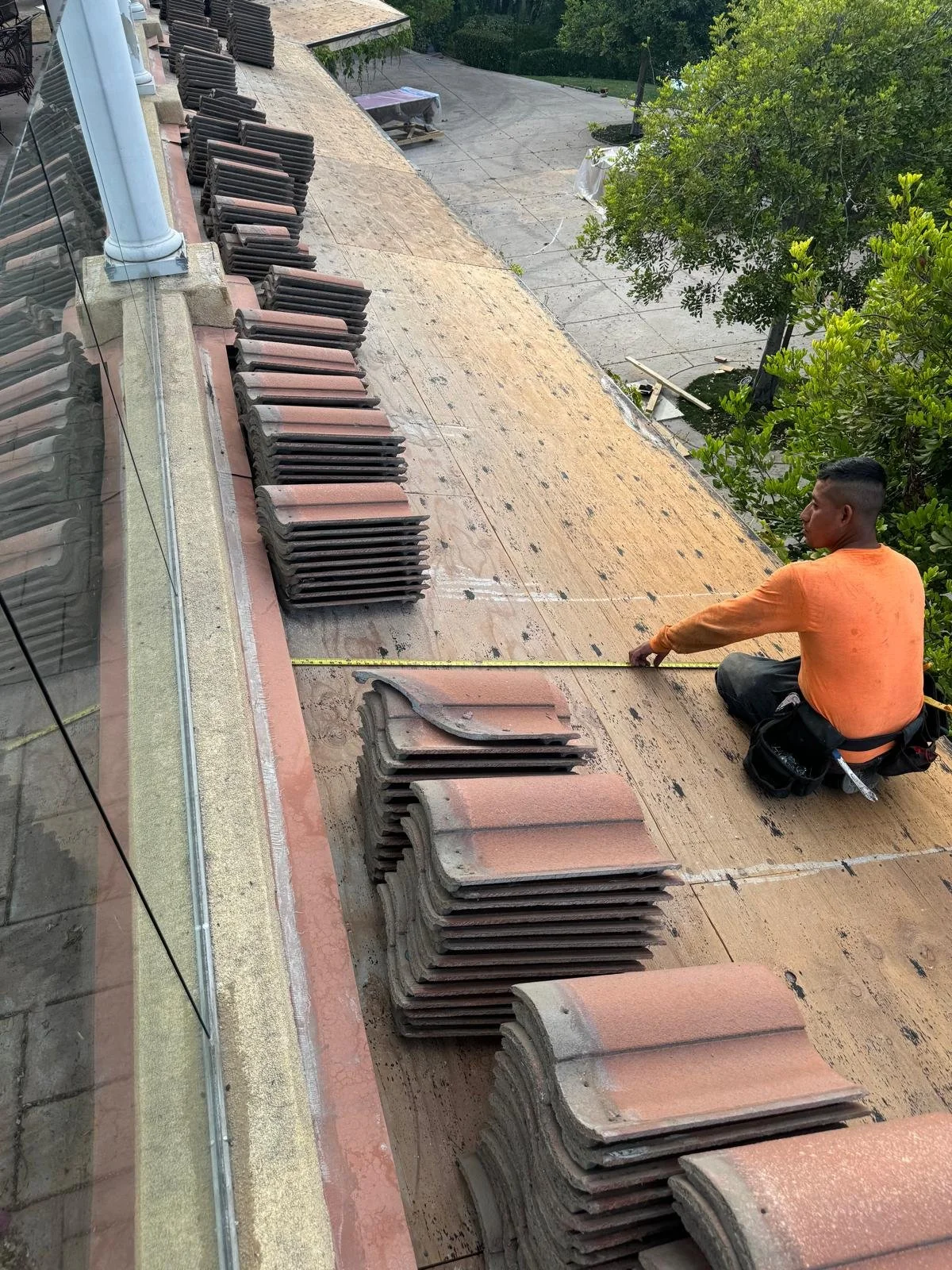 A worker in an orange shirt measures the width of a roof using a tape measure while sitting on the roof, with stacks of roof tiles around him and trees in the background.