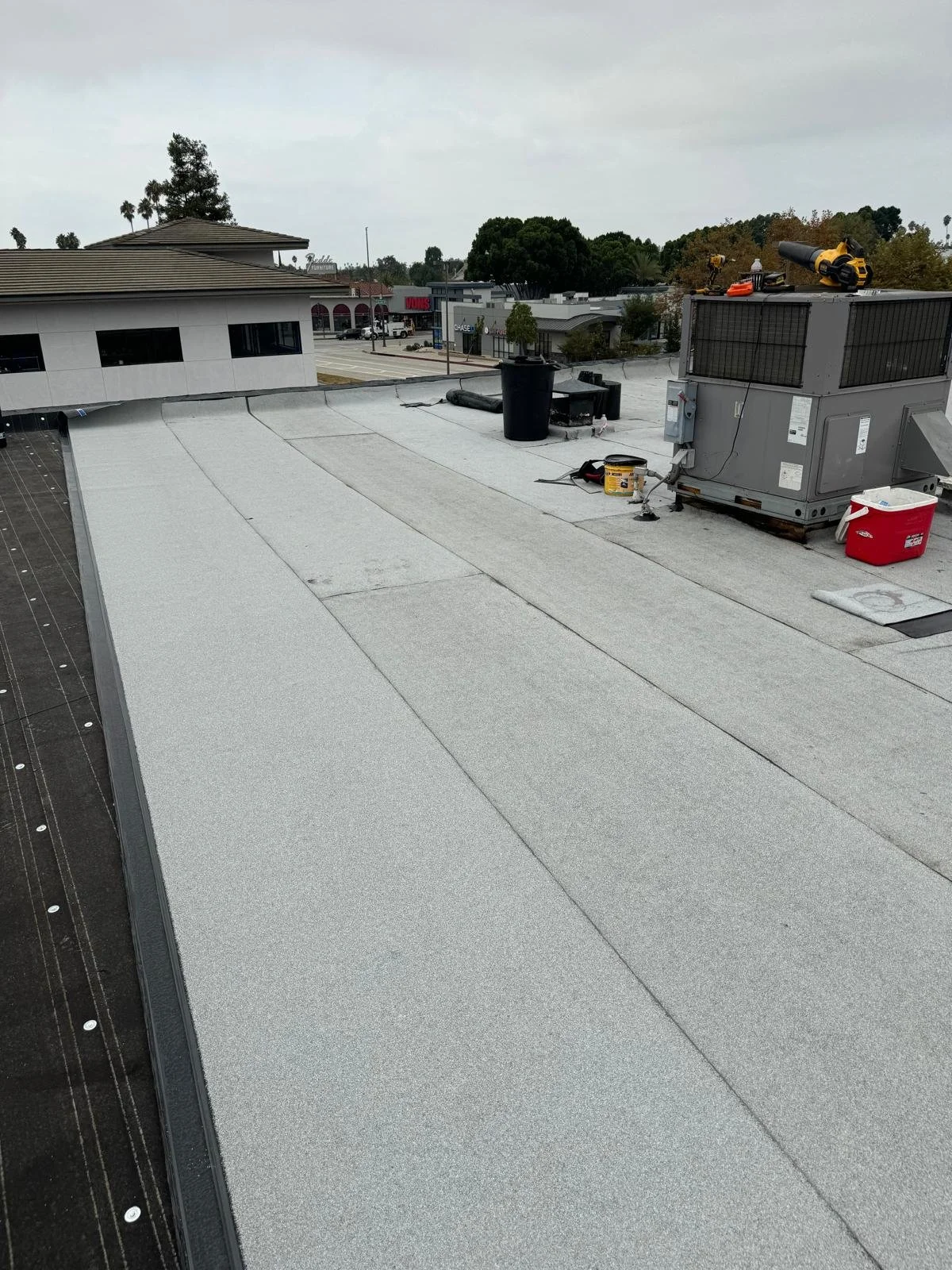 View of a commercial roof with light gray roofing material, HVAC unit, and tools on top, with a shopping area and trees visible in the background.