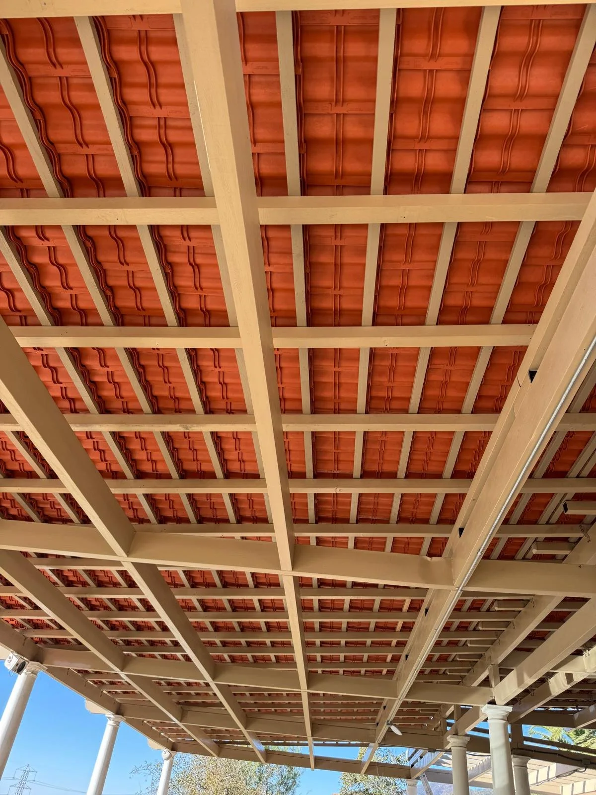 View of a wooden deck ceiling with exposed beams and red clay roof tiles, supported by white columns, with blue sky and trees visible in the background.