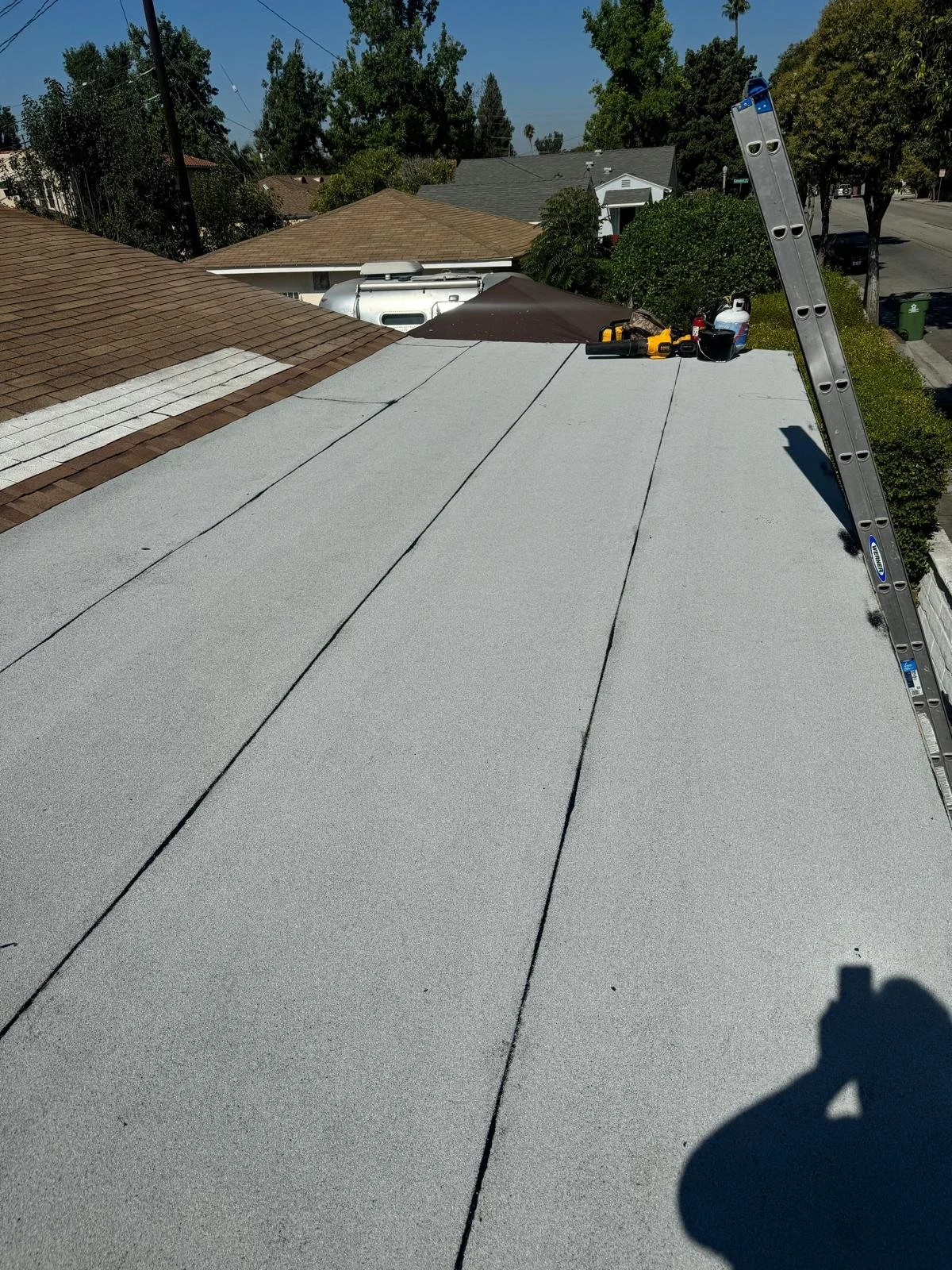 A rooftop with a section of new grey roofing material and tools, including a ladder leaning on the right side, and neighboring houses and trees in the background.