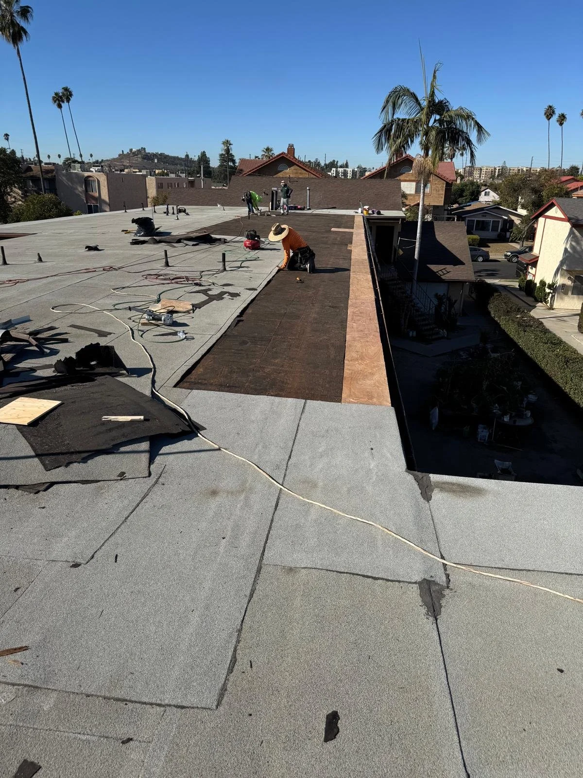 Workers on a flat rooftop performing construction or repair work with various tools and materials, under a clear blue sky.