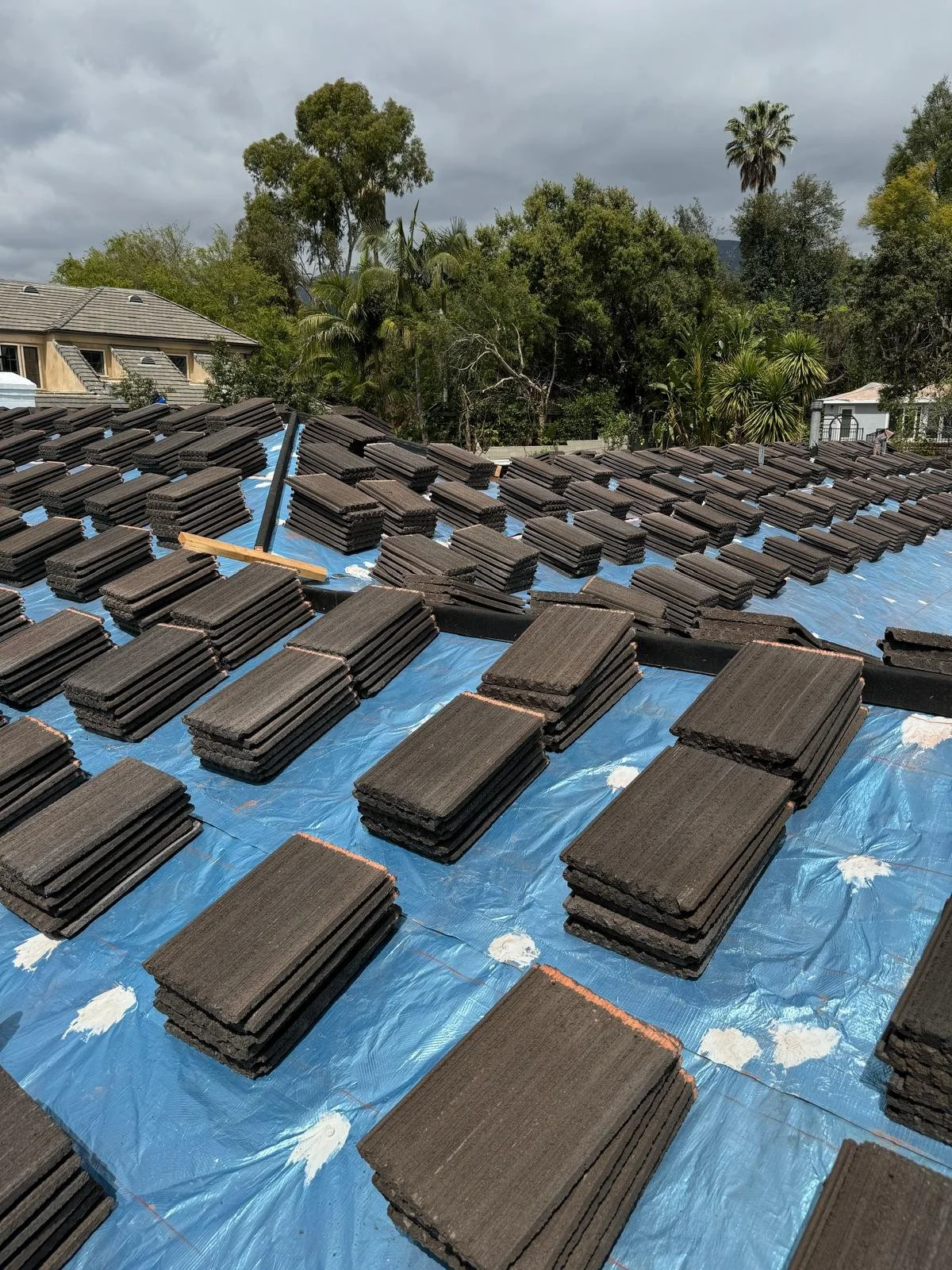 Roof being re-shingled with dark tiles, blue tarp underneath, and a ladder on the roof. House and trees in the background under cloudy sky.