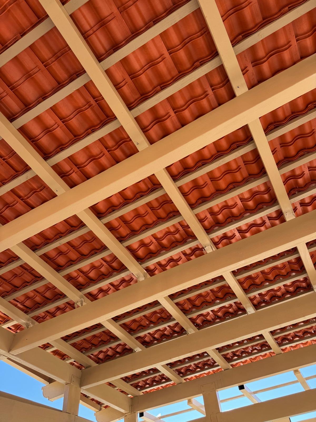 View of a roof structure with wooden beams and reddish-brown wavy roof tiles underneath, with a small part of a blue sky visible in the bottom left corner.