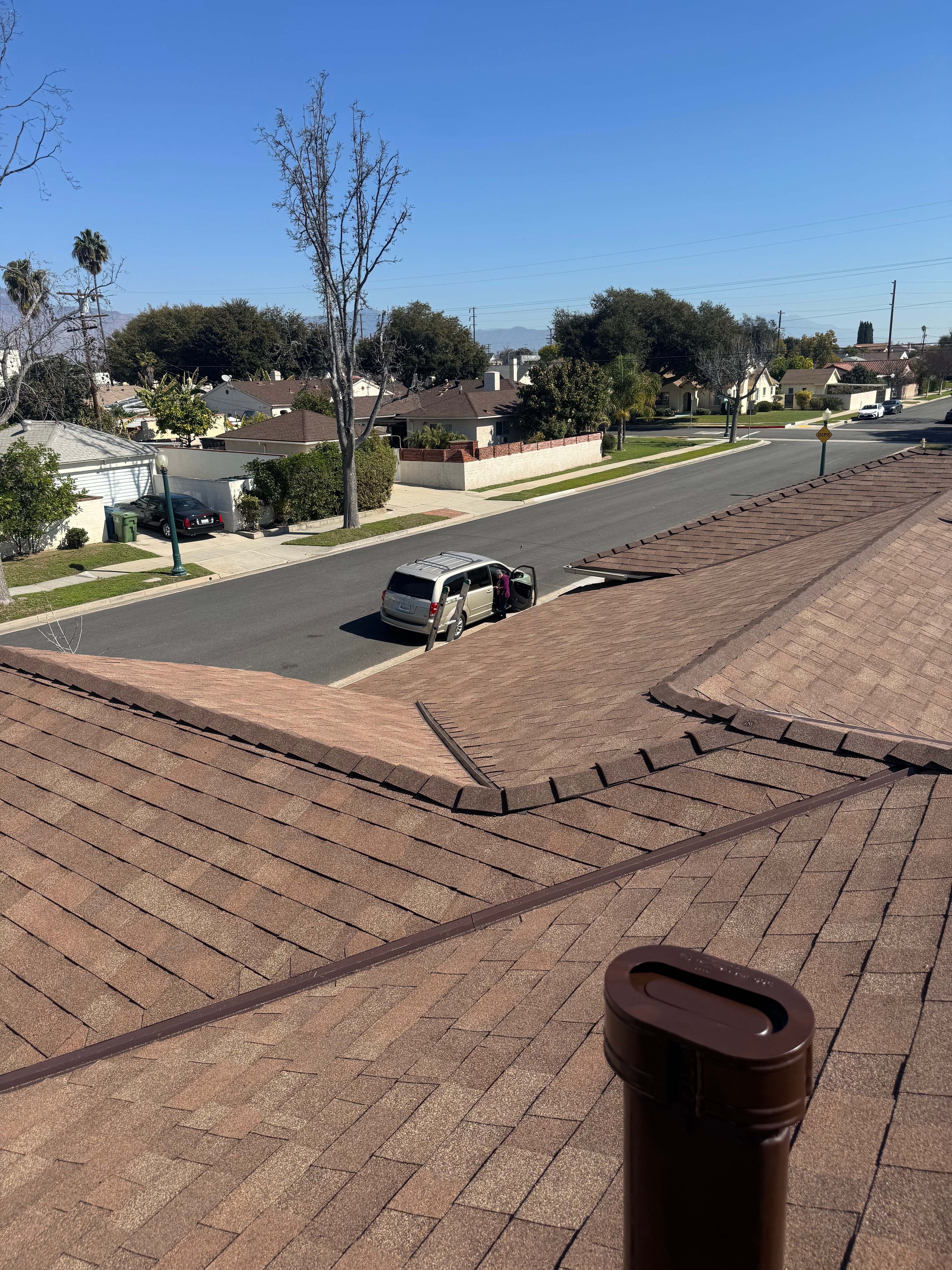 View from a rooftop showing neighboring houses, trees, a street with parked cars including an open minivan, and power lines under a clear blue sky.