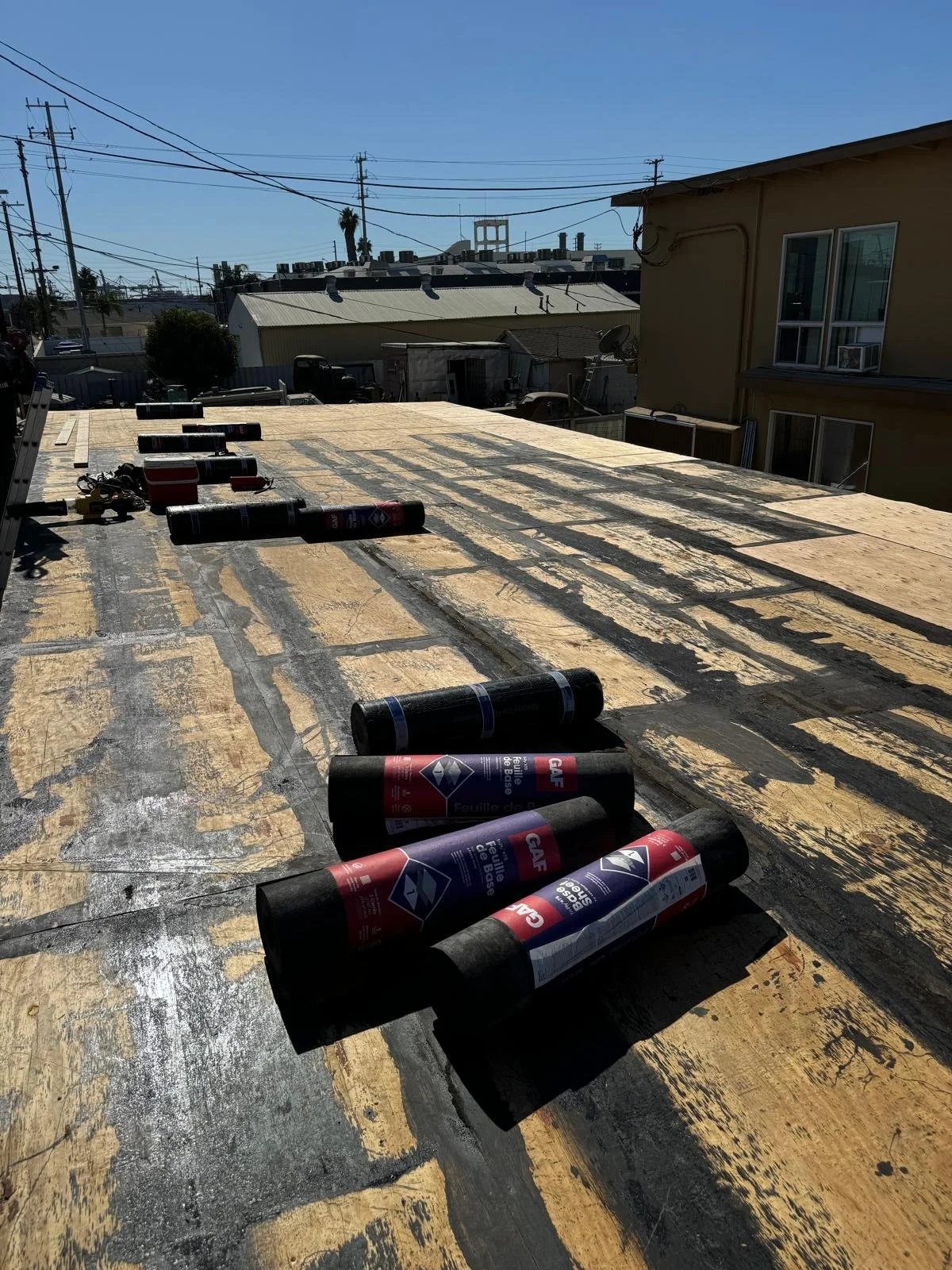 Rolls of roofing felt on a flat rooftop under a clear blue sky with neighboring buildings and utility poles in the background.