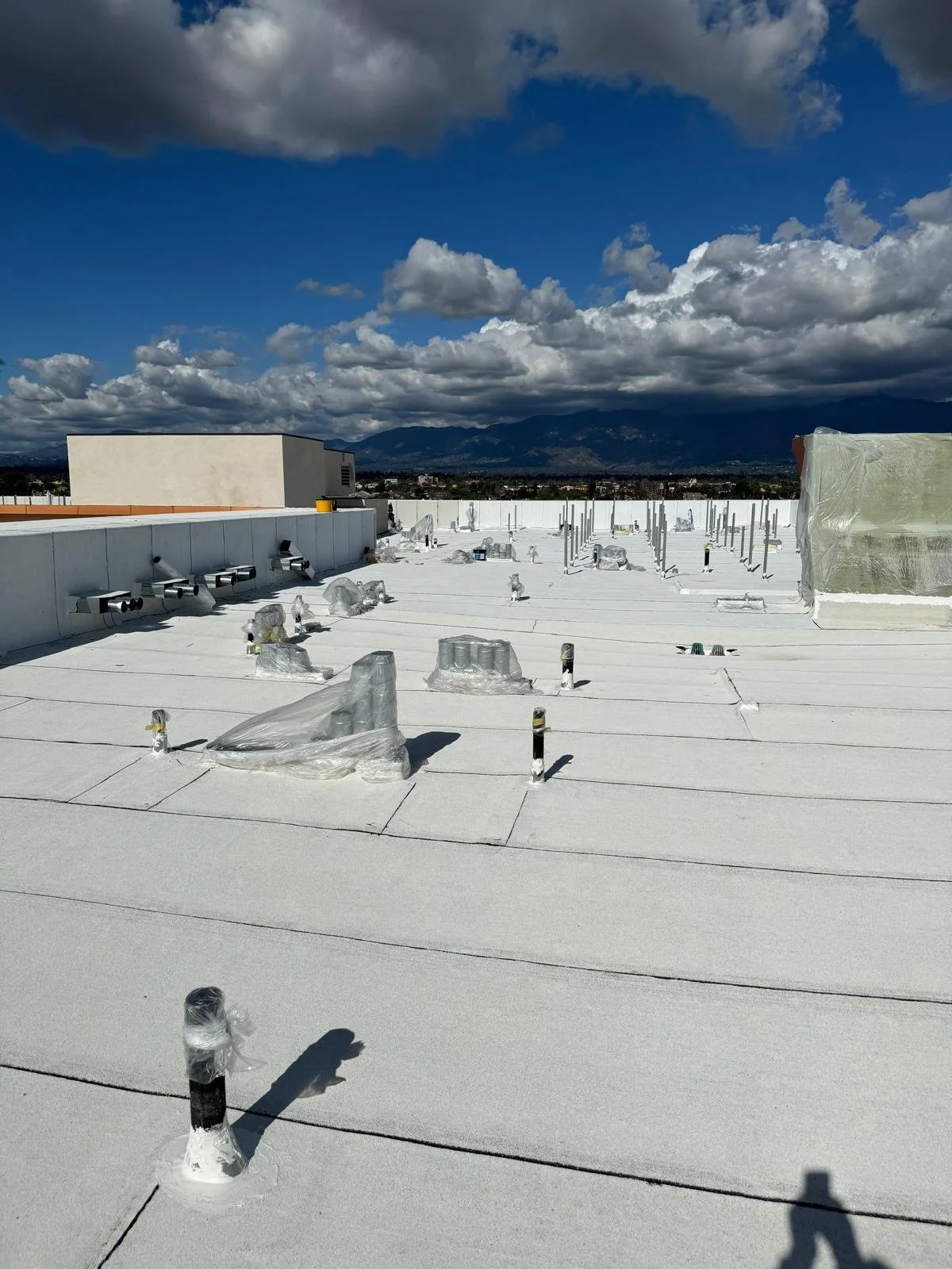 Rooftop with white waterproof membrane, several vents and pipes wrapped in plastic, under a partly cloudy sky, with mountains visible in the distance.
