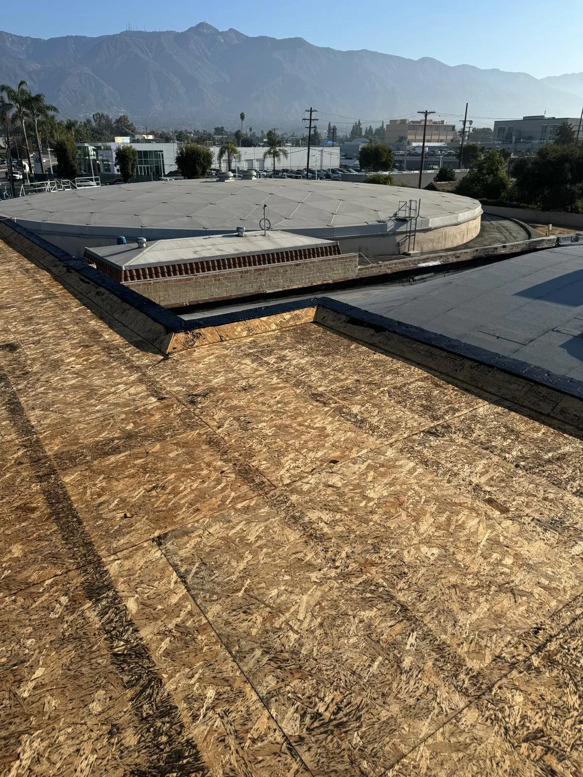 View from a rooftop showing a flat building roof with OSB sheathing, in the background there are other rooftops, trees, power poles, and mountains under a clear sky.