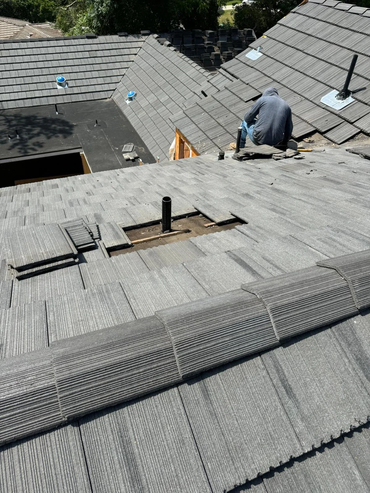A person working on the roof of a house, installing or repairing gray roofing shingles, with some shingles stacked nearby.