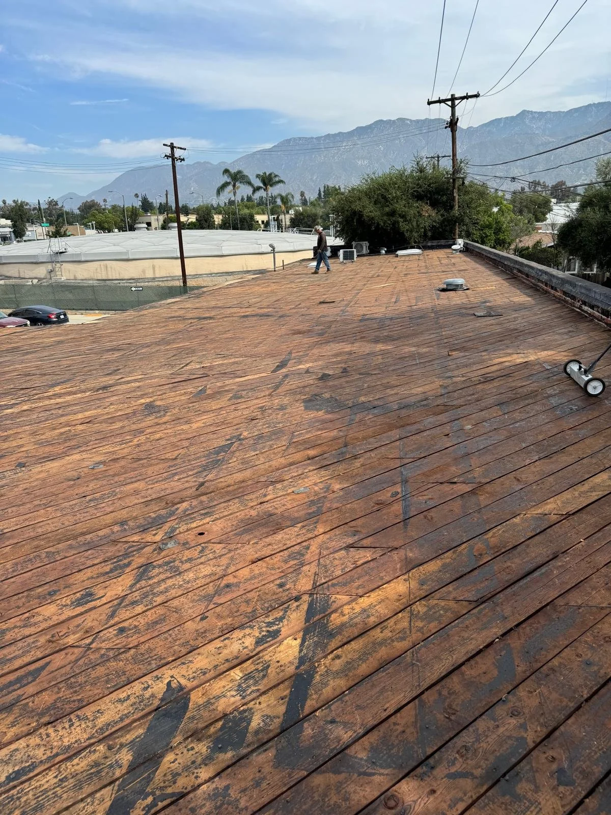 A view of a rooftop with weathered wooden planks, power lines, and a person walking in the distance against a background of trees, mountains, and a partly cloudy sky.