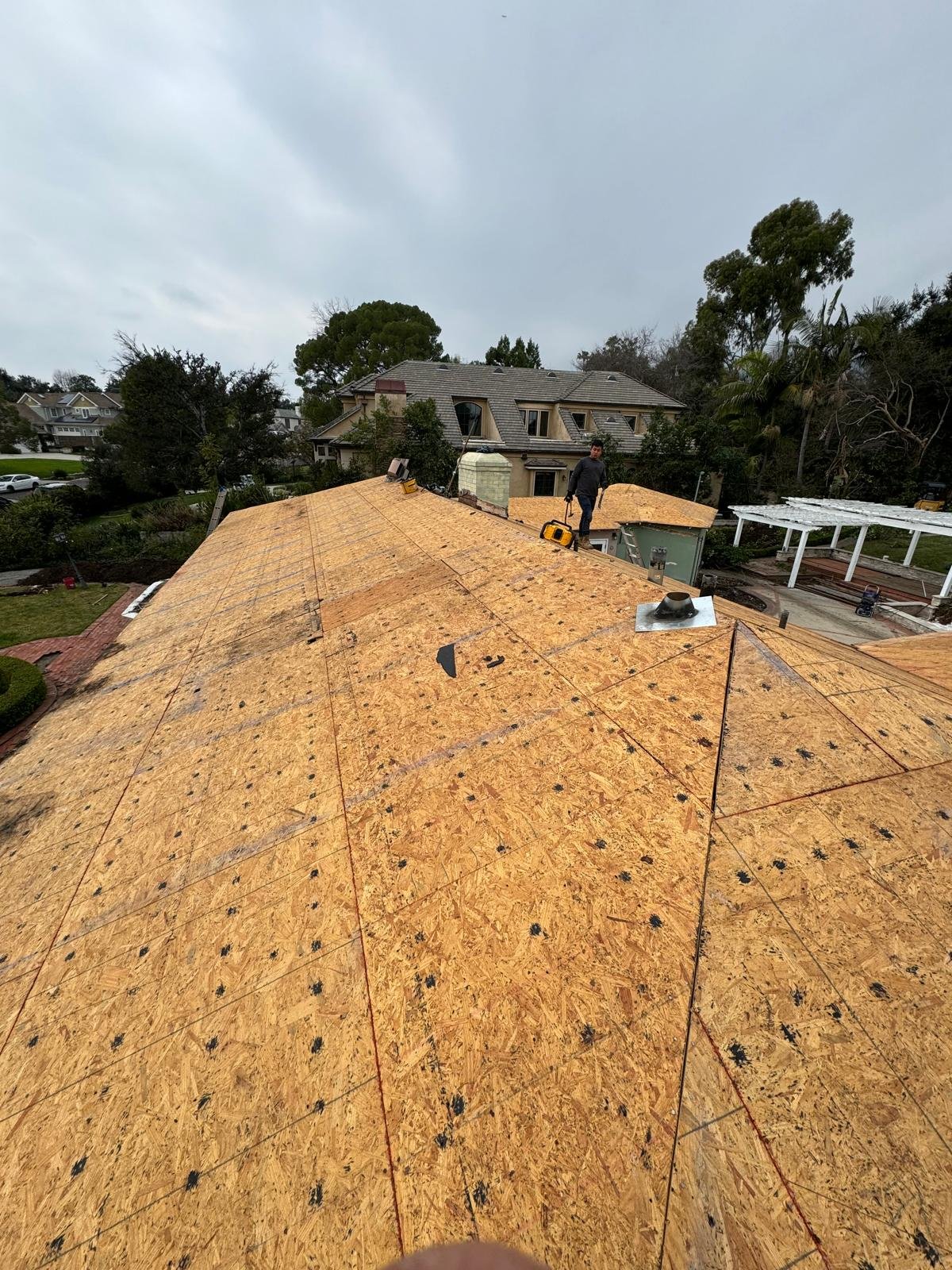 A construction worker standing on a wooden roof structure under an overcast sky, with residential houses and trees in the background.
