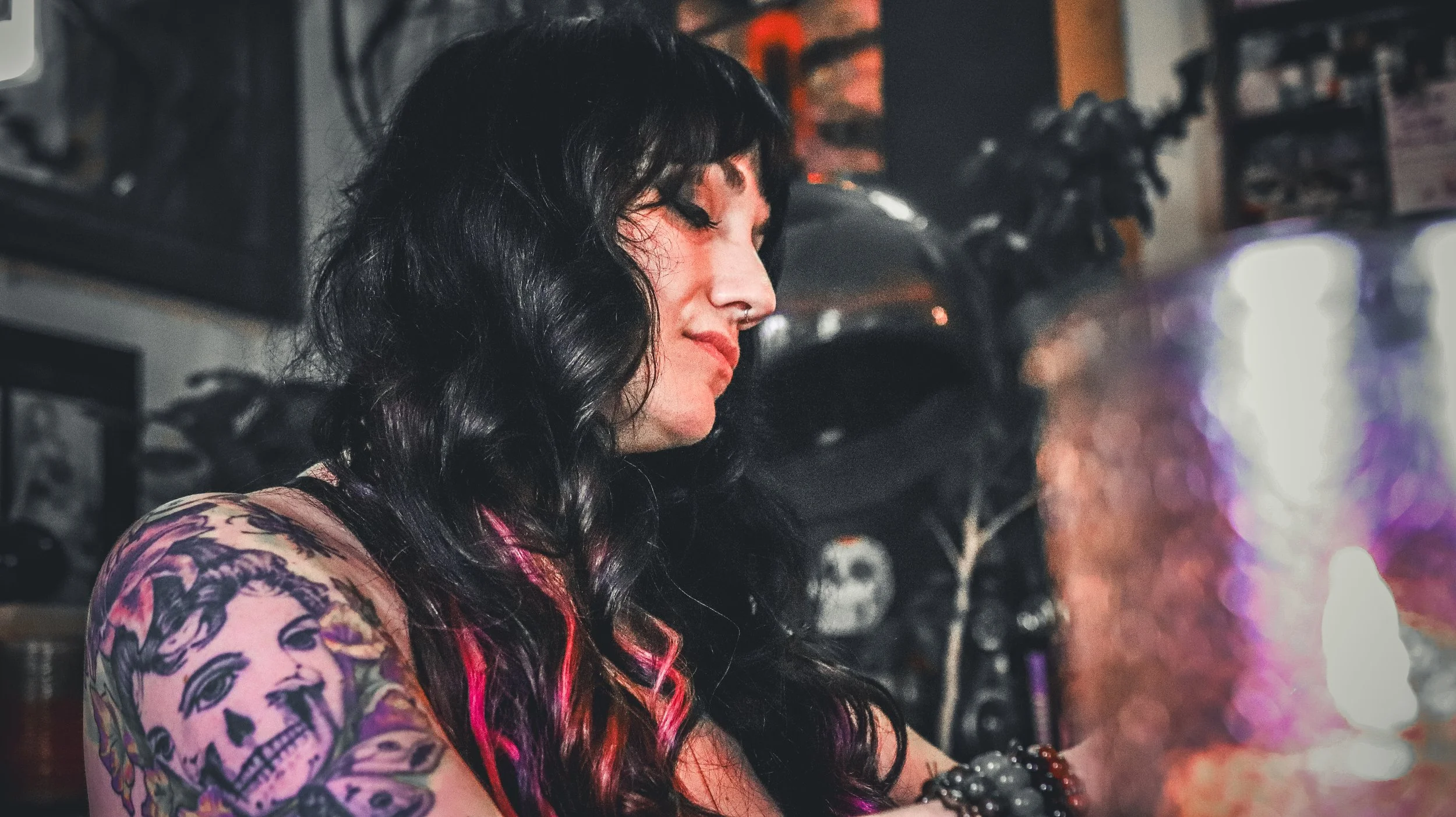 A woman with black wavy hair, tattoos on her arm, and a septum piercing is sitting in a dimly lit room, looking downward with a contemplative expression. The background features motorcycle helmets, a skull, and shelves, with some colorful lights.