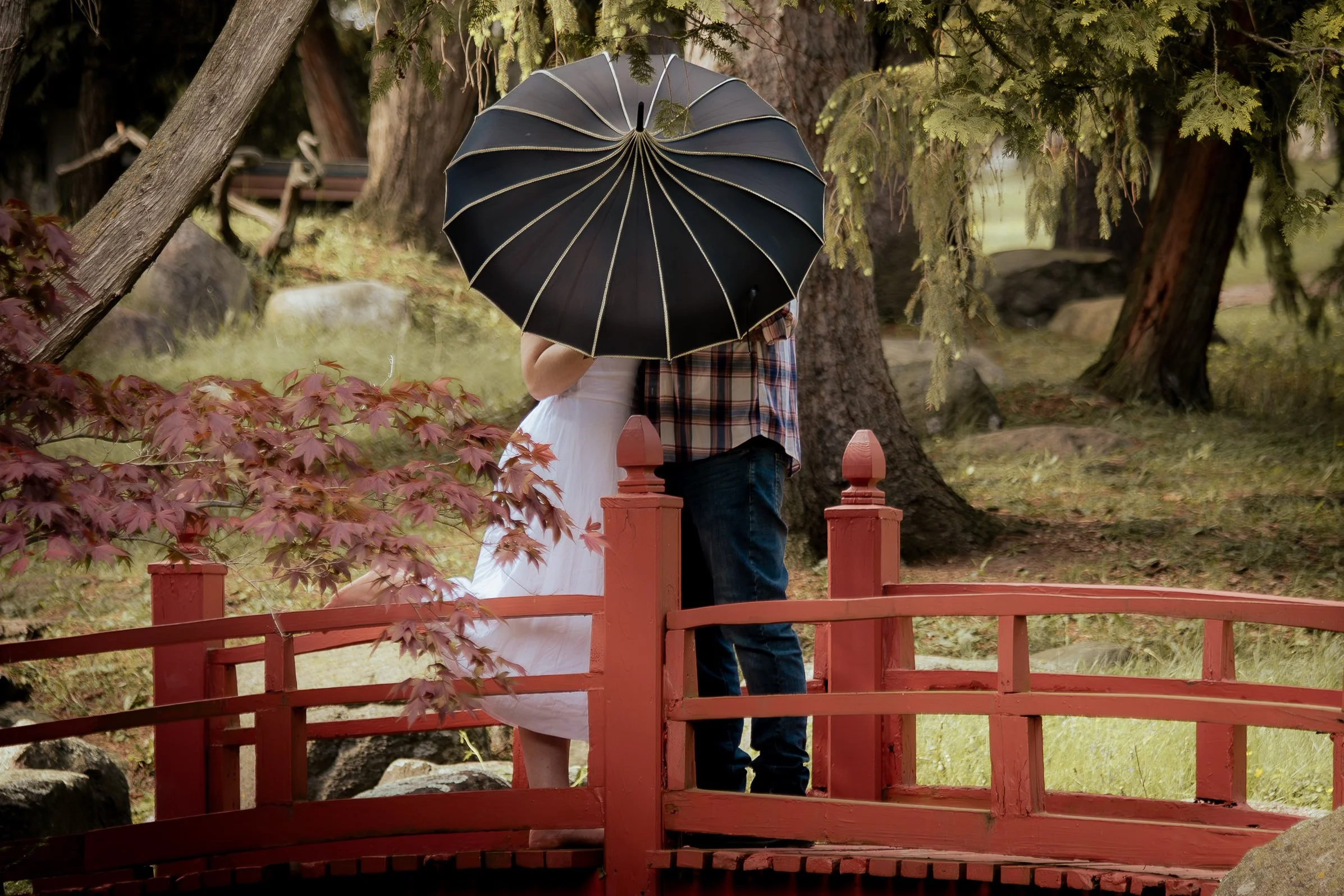 A couple standing on a red wooden bridge holding a large black umbrella in a park with trees and purple leaves.