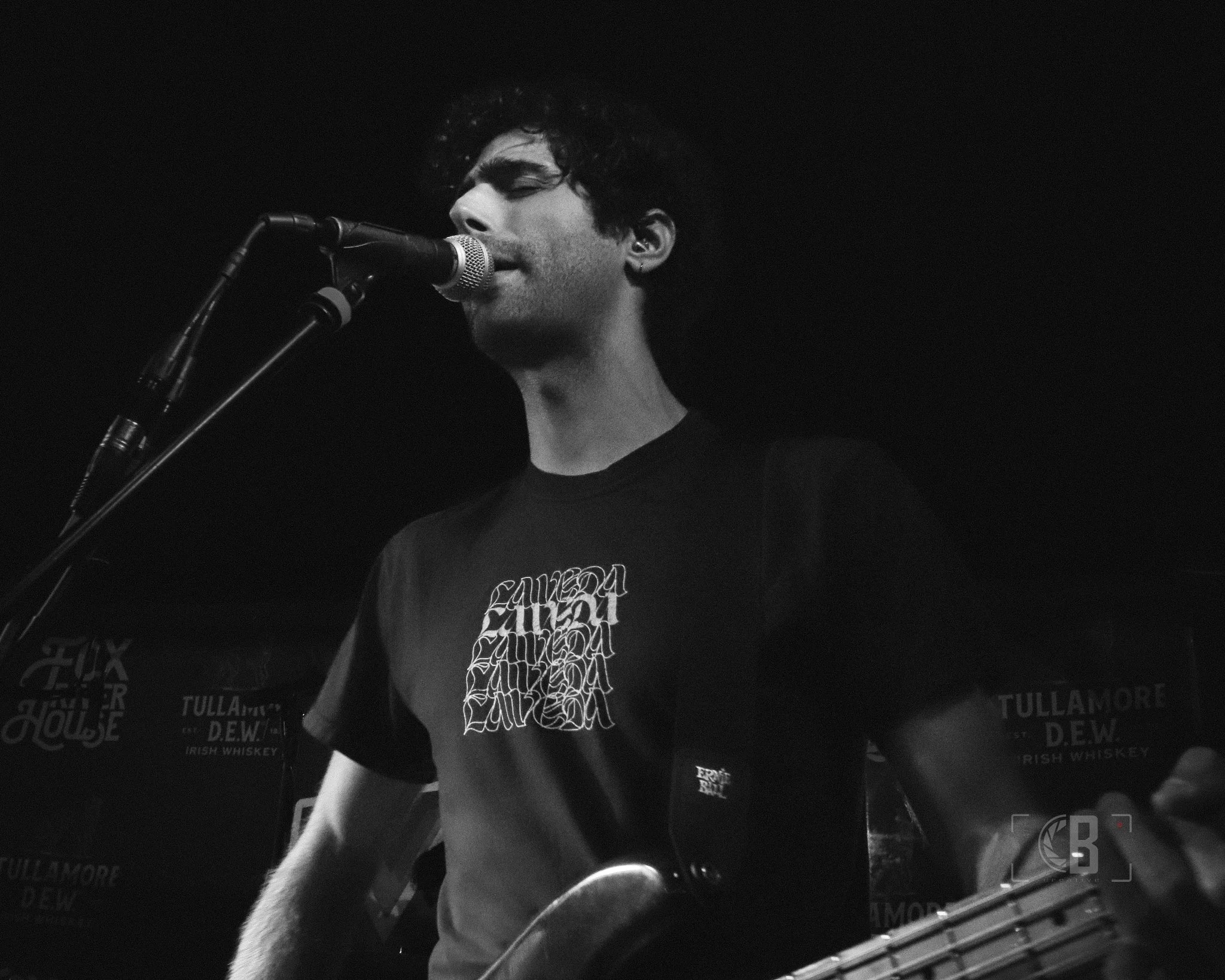 A black-and-white photo of a man singing into a microphone while playing a bass guitar during a live performance.