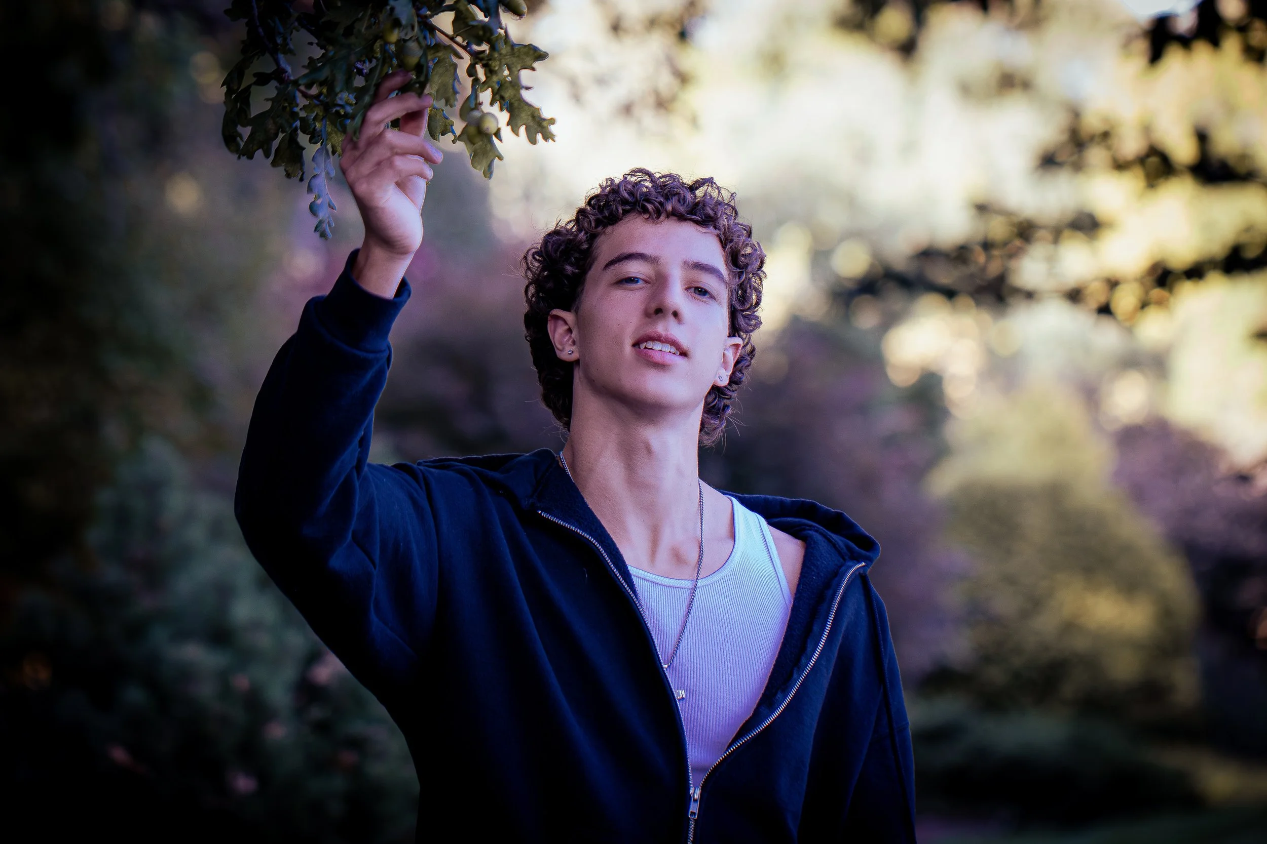 A young person with curly hair and earrings, wearing a striped t-shirt and a black hoodie, stands outdoors touching a leafy tree branch with small green fruit, with blurred trees and sunlight in the background.