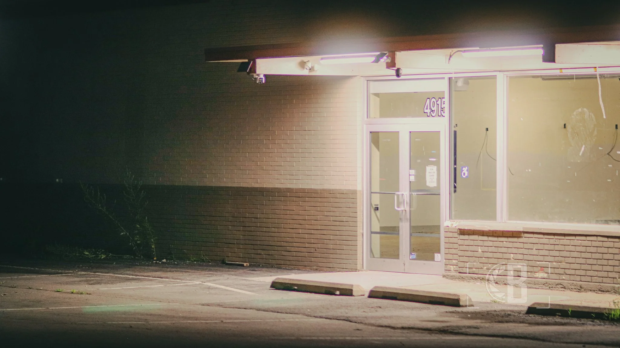An empty brick storefront with glass doors and windows, illuminated by bright fluorescent lighting, with parking spaces in front and minimal surroundings.